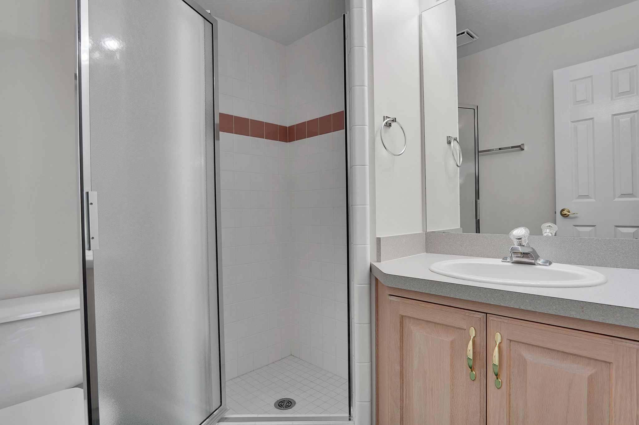 Bathroom featuring a glass-enclosed shower, white tiled walls with a decorative accent band, a vanity with a light-colored countertop, and wood-finish cabinetry with brass-tone hardware