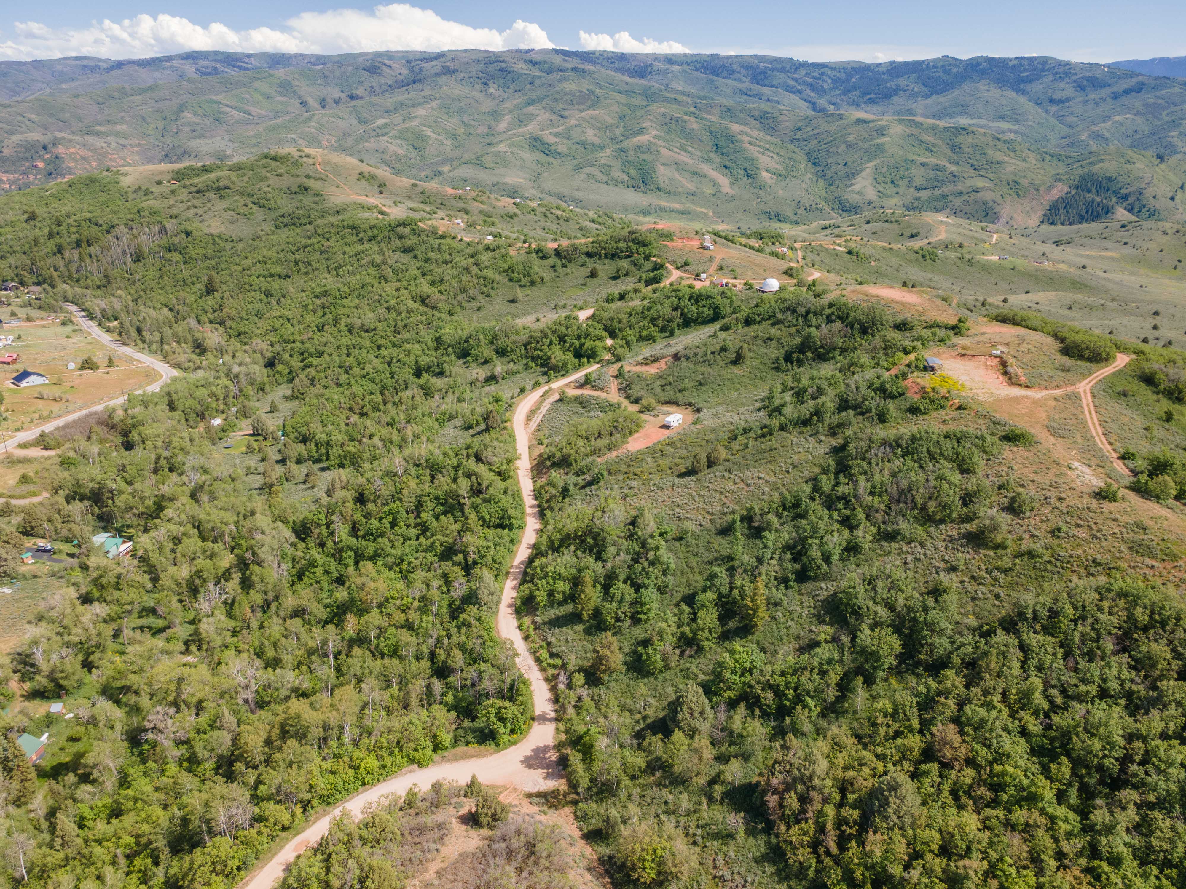 Expansive mountainous terrain featuring a winding unpaved road, scattered green trees, and distant natural hills