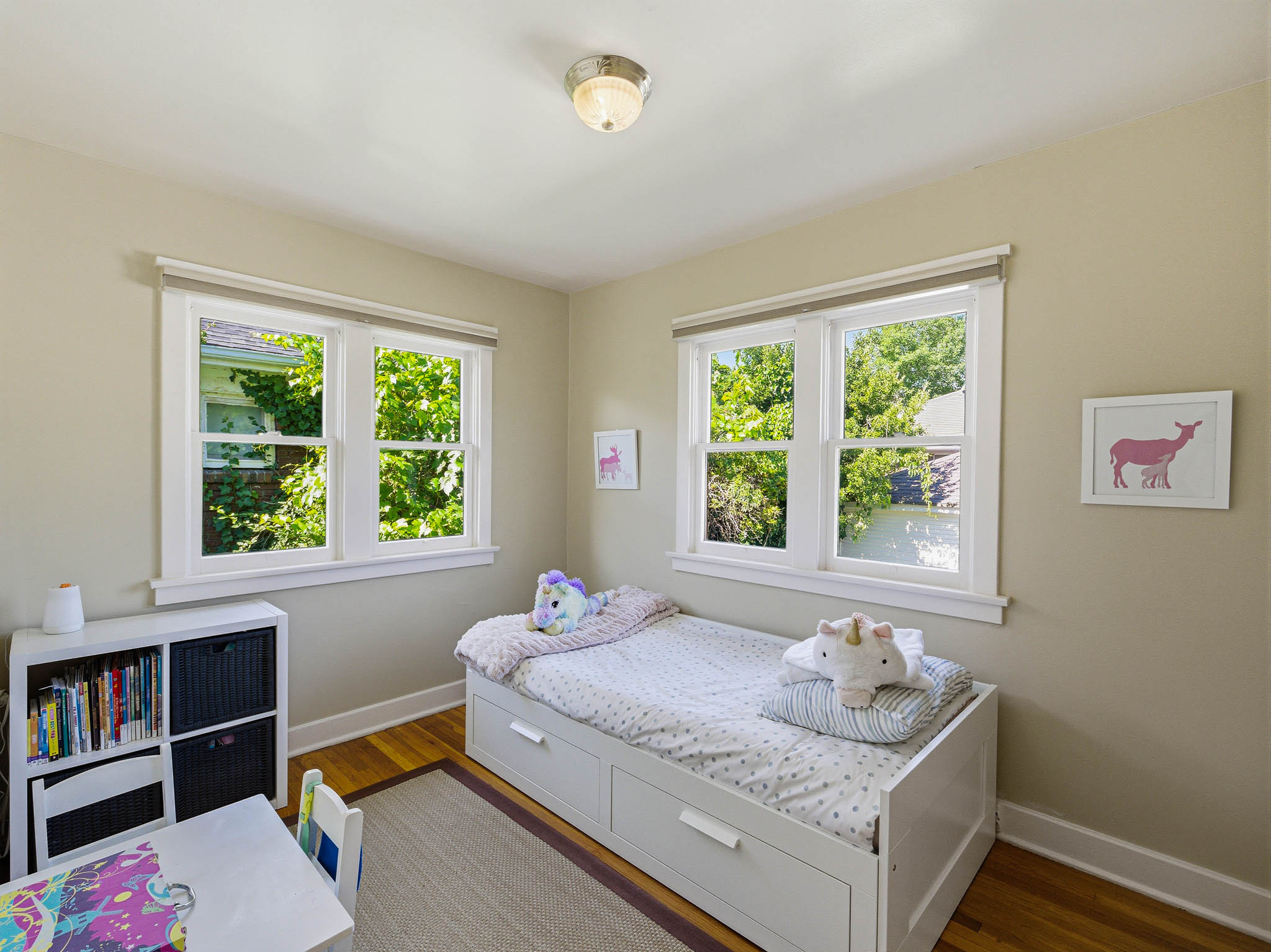 Hardwood flooring throughout this room, featuring two large windows with white trim, light-toned walls, and a flush-mount ceiling light fixture
