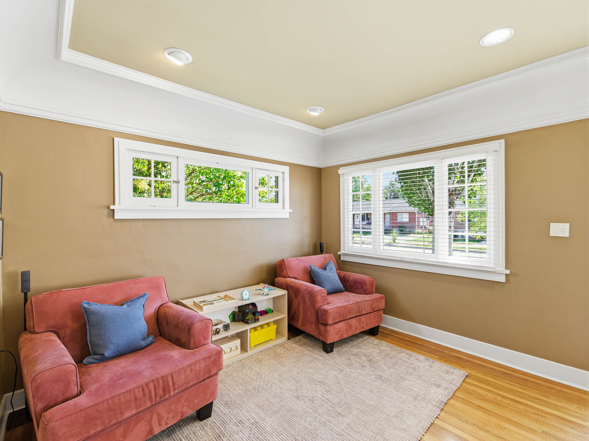 Recessed ceiling lighting illuminates a room featuring polished wood-finish flooring, a tray ceiling with white crown molding, and two windows with white trim