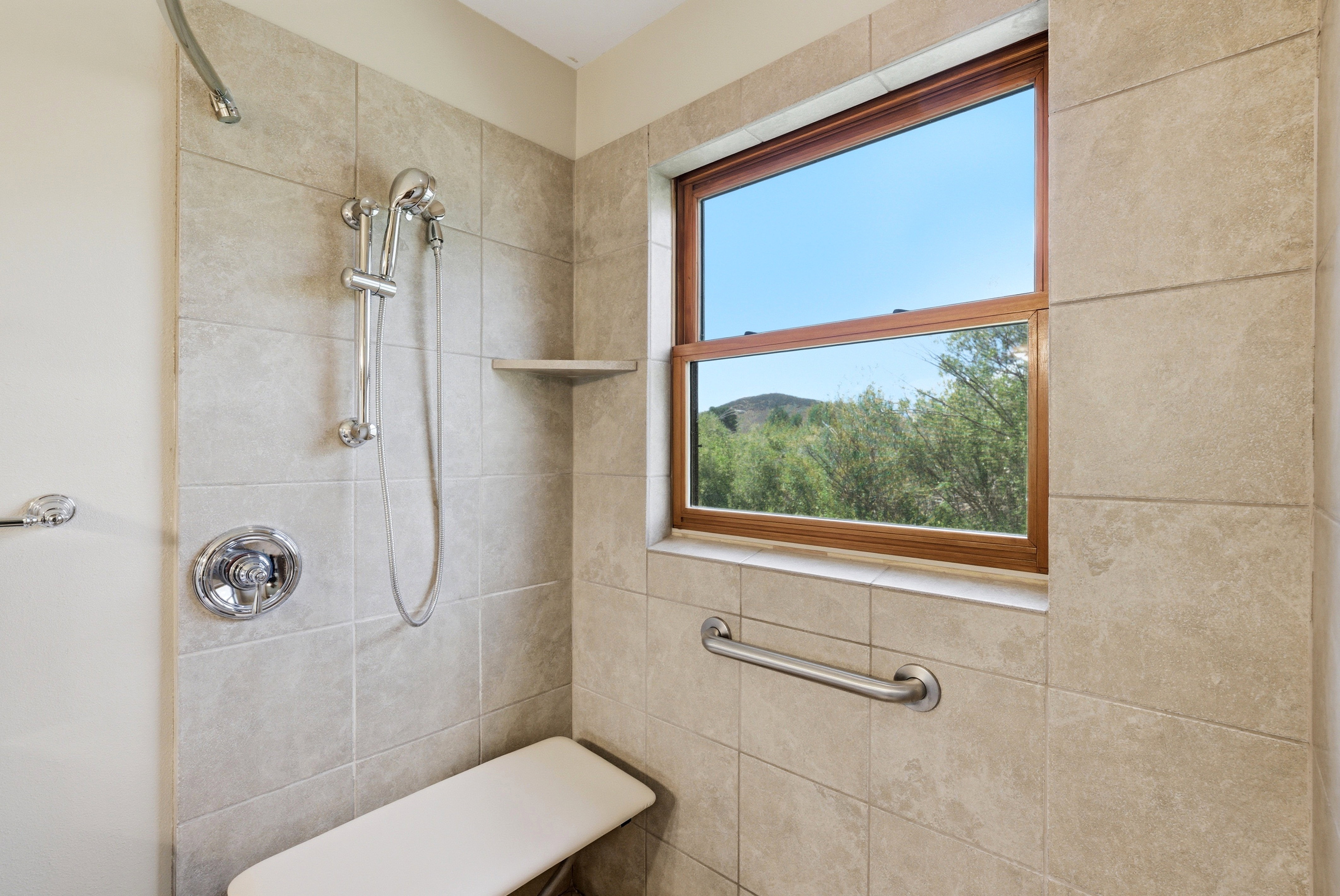 Tiled shower enclosure featuring a wood-framed window, chrome fixtures, a handheld showerhead, an integrated shower bench, and a grab bar