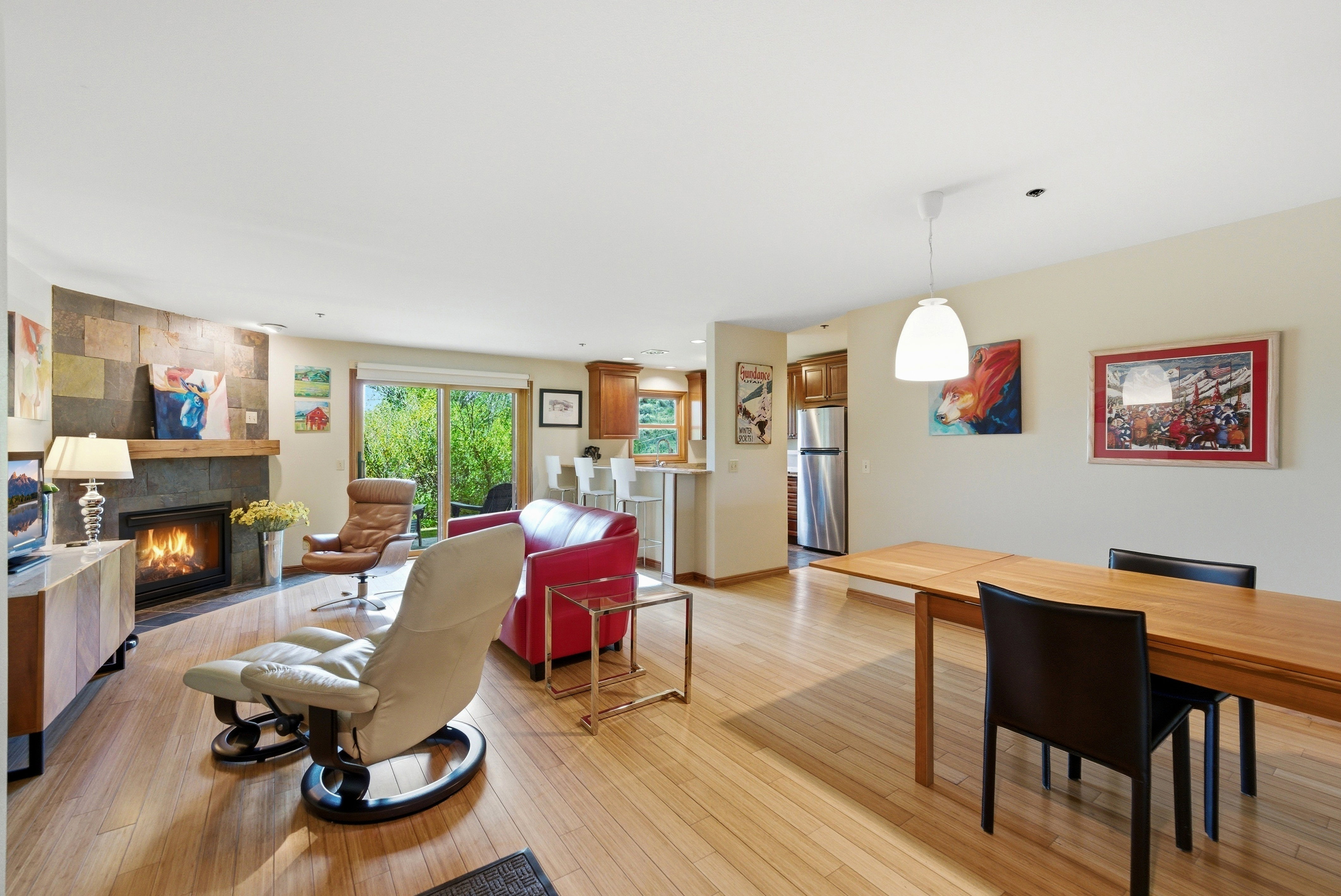 Open-concept living space featuring wood-finish flooring, a stone-accented fireplace with a wood mantel, a sliding glass door for exterior access, a built-in kitchen island with seating, and a dining area with pendant lighting
