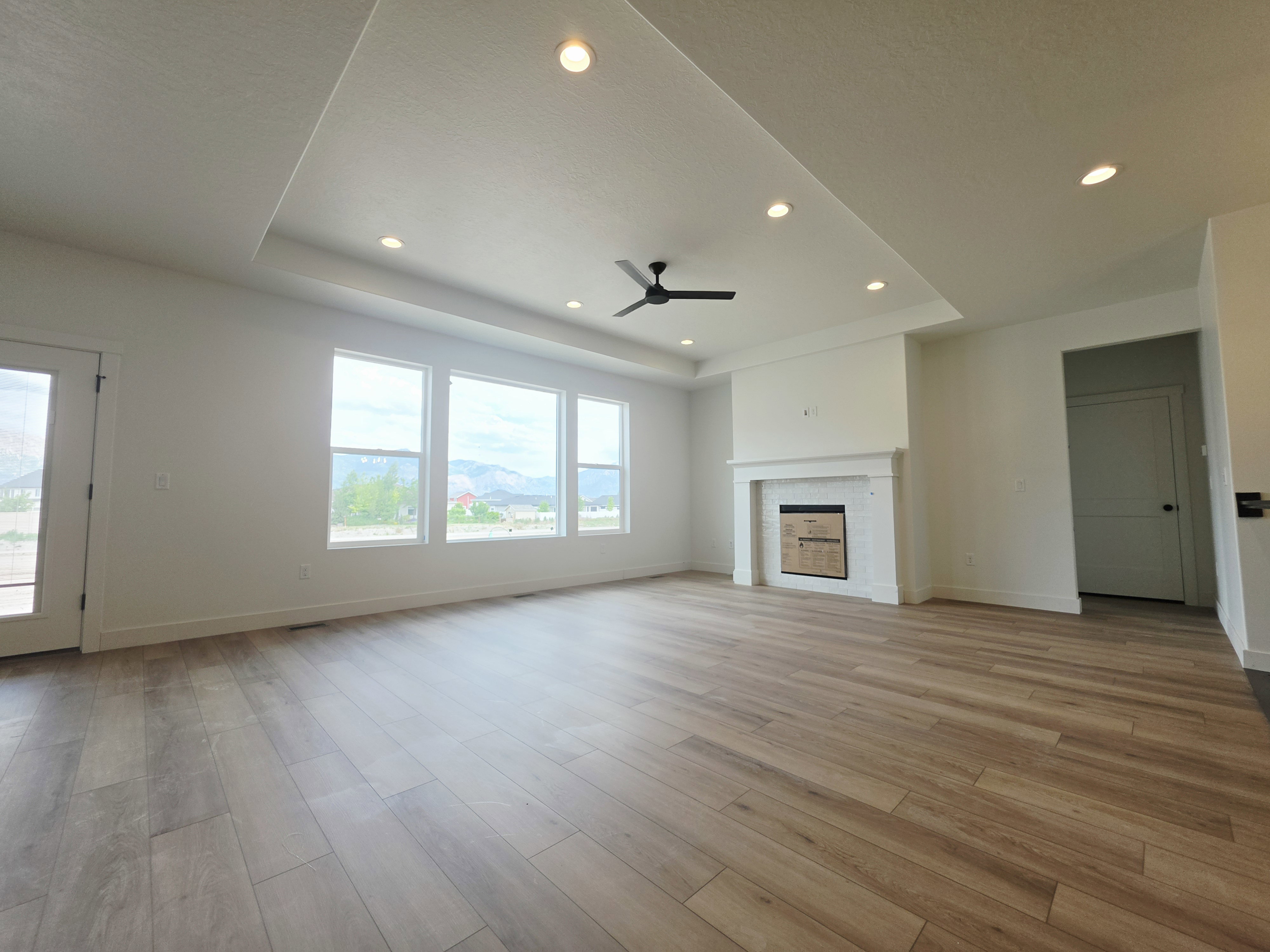 Spacious interior featuring wood-finish flooring, a tray ceiling with recessed lighting and a ceiling fan, a fireplace with a white mantel and tile surround, and multiple windows providing natural light