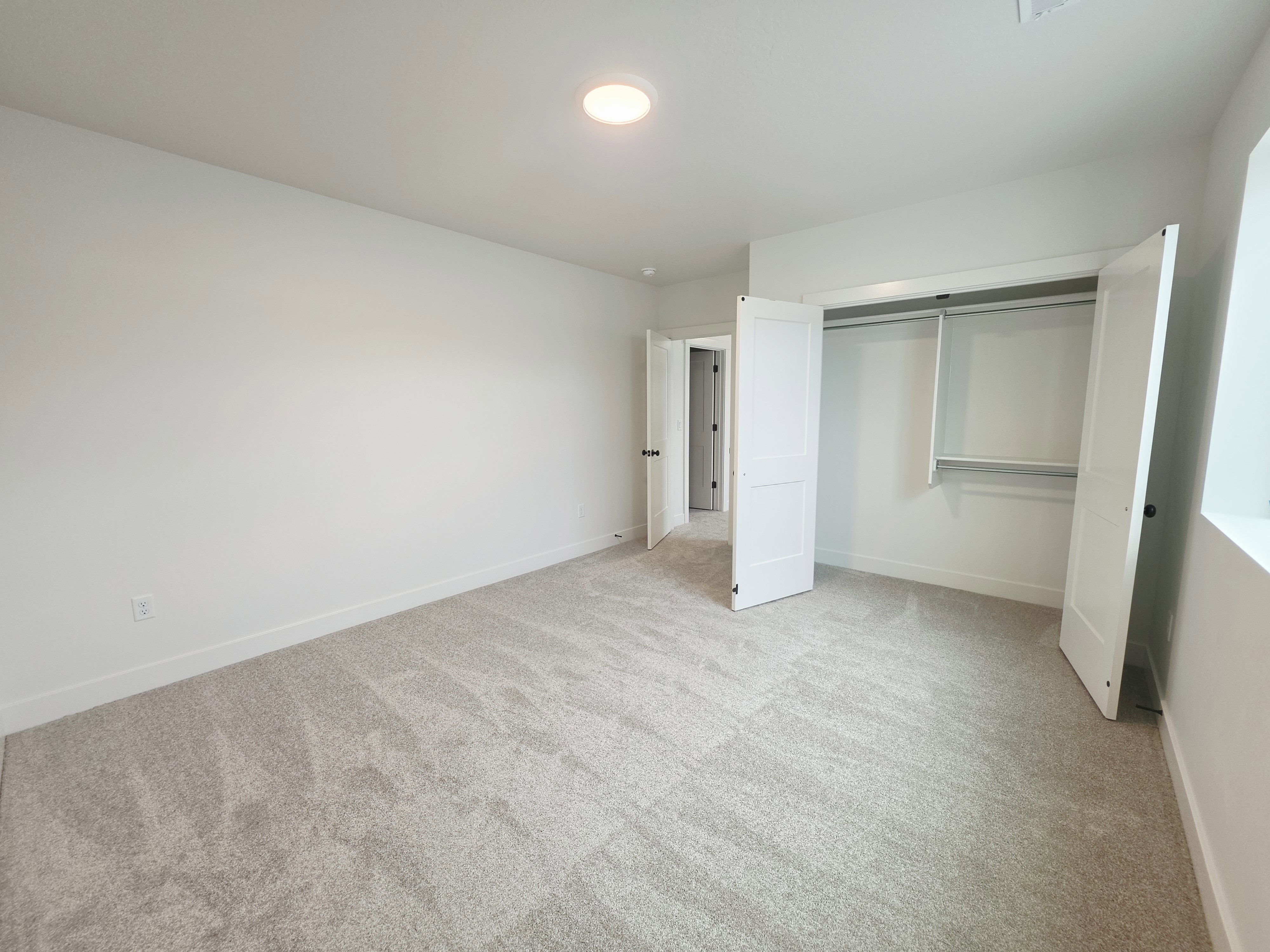 Spacious room featuring light gray carpeting, white baseboards, a flush-mount ceiling light, and a double-door closet with shelving and hanging rods