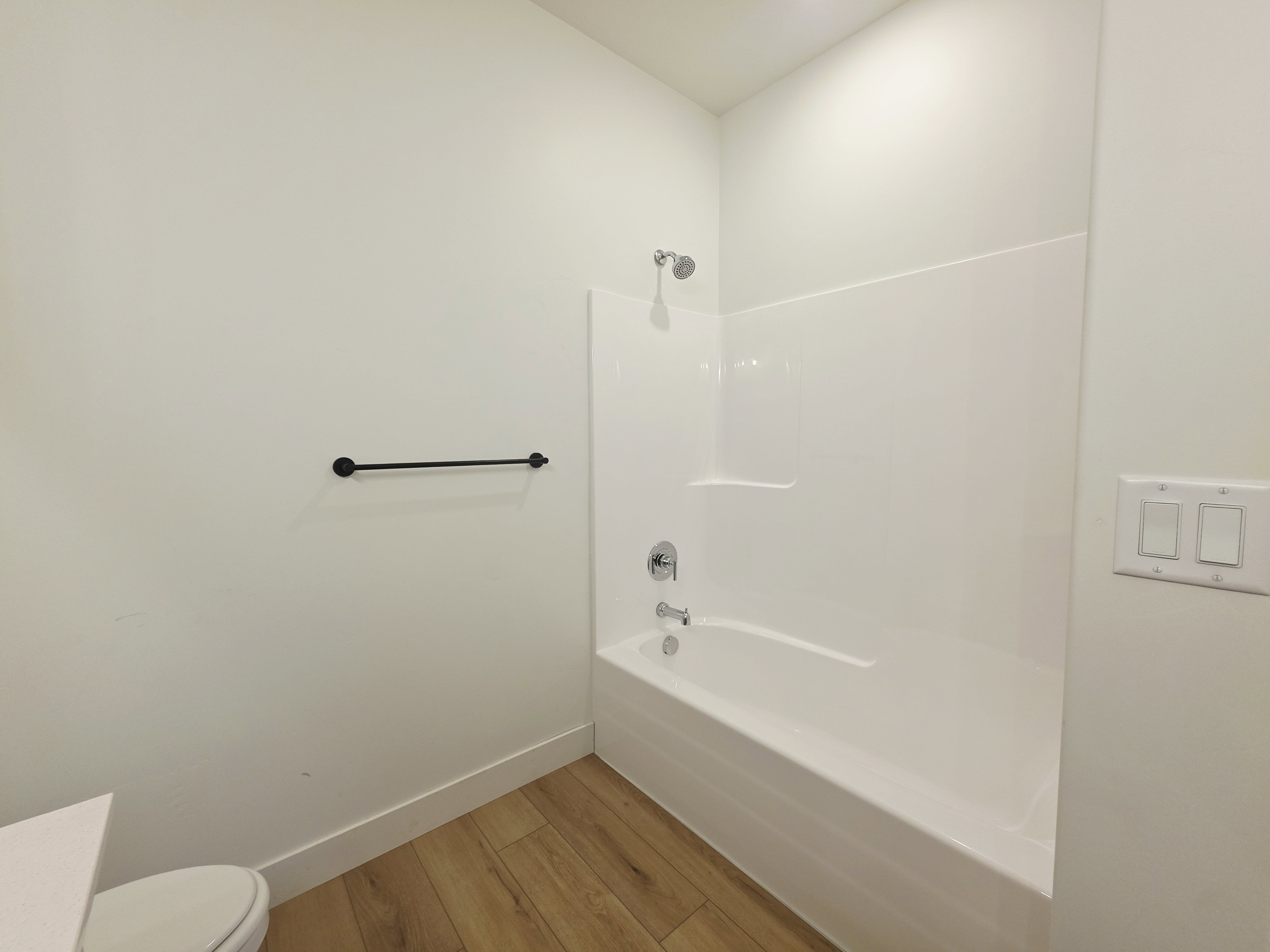 Bathroom featuring a white tub/shower combination with chrome fixtures, a modern black towel bar, light wood-finish flooring, and crisp white walls