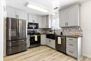 Contemporary kitchen featuring light gray cabinetry with black hardware, stainless steel appliances, wood-finish flooring, a black farmhouse sink, and mosaic tile backsplash
