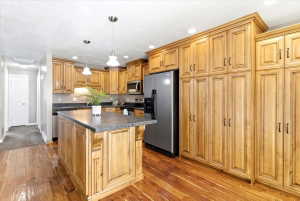 Kitchen featuring natural wood cabinetry, dark countertops, stainless steel appliances, a central island with pendant lighting, and wood-finish flooring