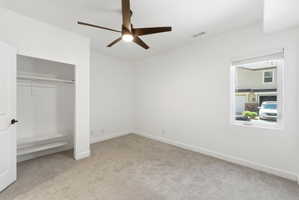Bright room featuring neutral carpeting, white walls, a modern ceiling fan with lighting, a window with roller blinds, and an open closet with built-in shelving and a clothing rod