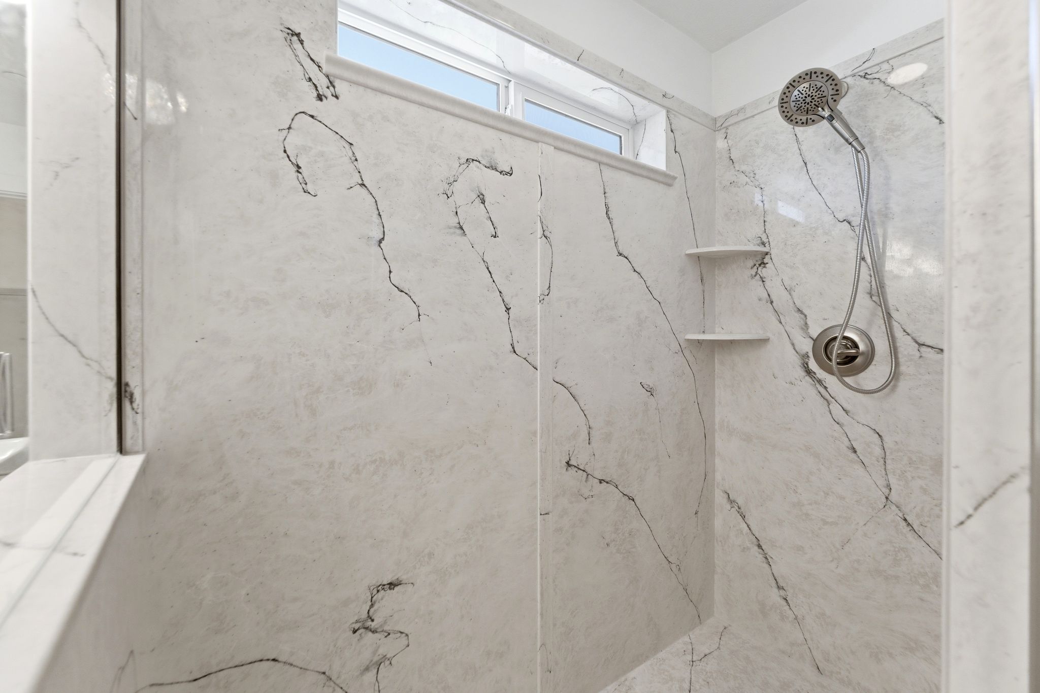 Shower stall featuring large-format wall panels with black veining, a multi-function showerhead, and integrated corner shelving