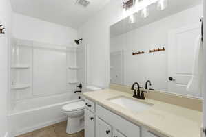 Full bathroom featuring a white tub/shower combination with integrated shelving, a white toilet, and a vanity with a light-toned countertop, integrated sink, and dark bronze-finish fixtures