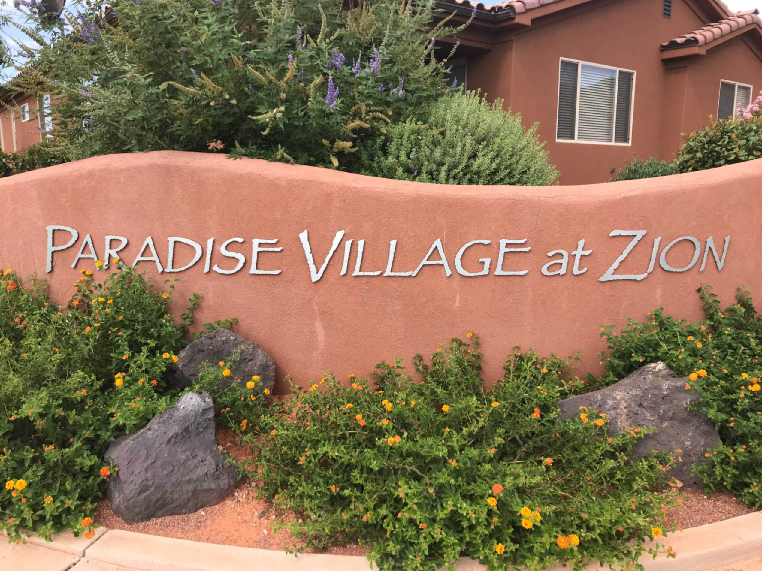 Stucco entrance monument featuring "PARADISE VILLAGE at ZION" lettering, surrounded by varied desert landscaping