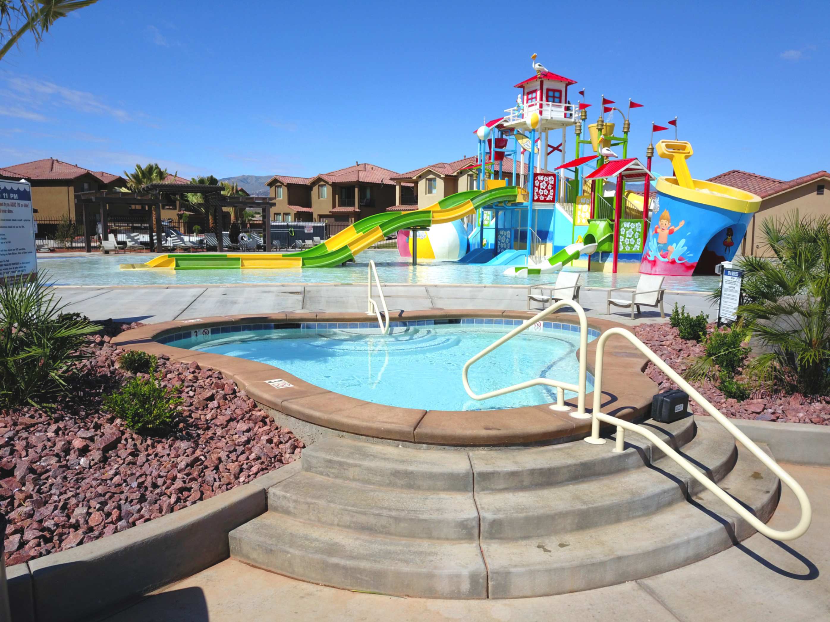 Resort-style aquatic amenity featuring a spa with integrated steps and handrails, a surrounding rock and shrub landscape, an expansive pool, and a colorful water play structure with multiple slides