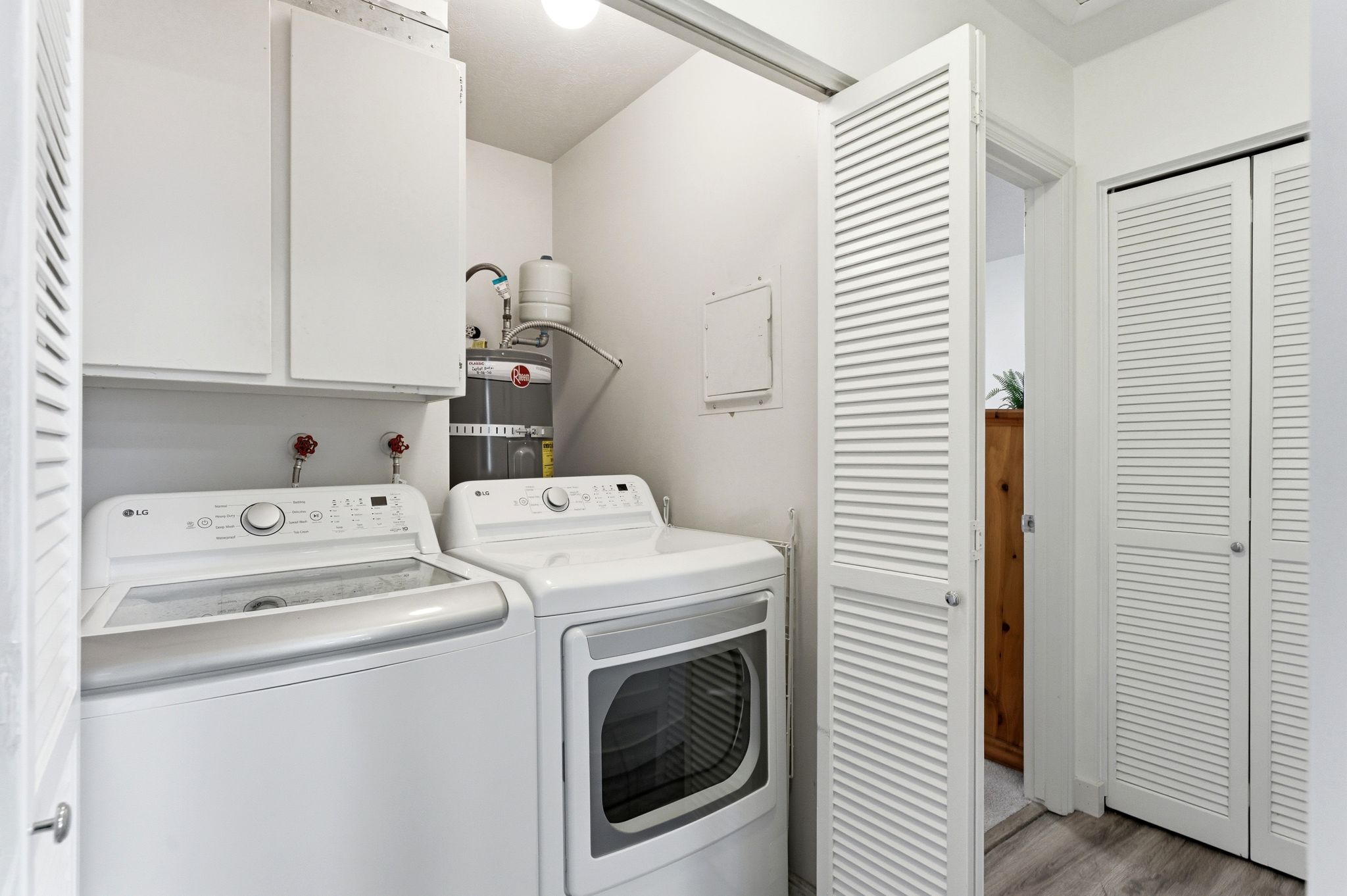 Dedicated utility closet with white louvered bifold doors, overhead cabinetry, and wood-finish flooring