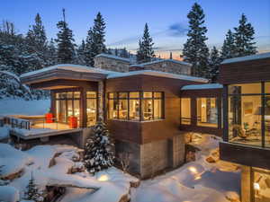 Modern mountain residence featuring extensive window walls, multi-level wood-clad exteriors, stone masonry accents, a suspended walkway, and a concrete foundational base