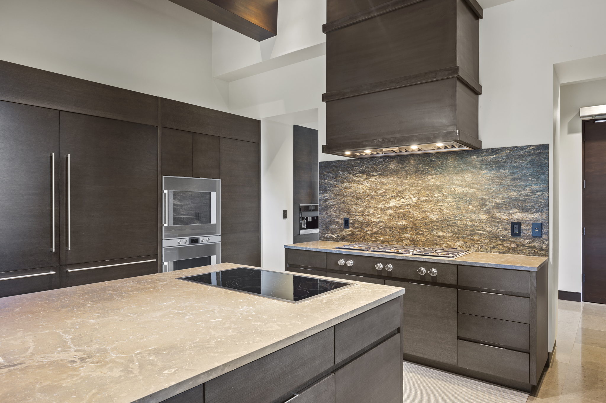 Kitchen featuring a large island with an integrated cooktop, dark wood-finish cabinetry, built-in stainless steel appliances, a stone backsplash, and a custom range hood
