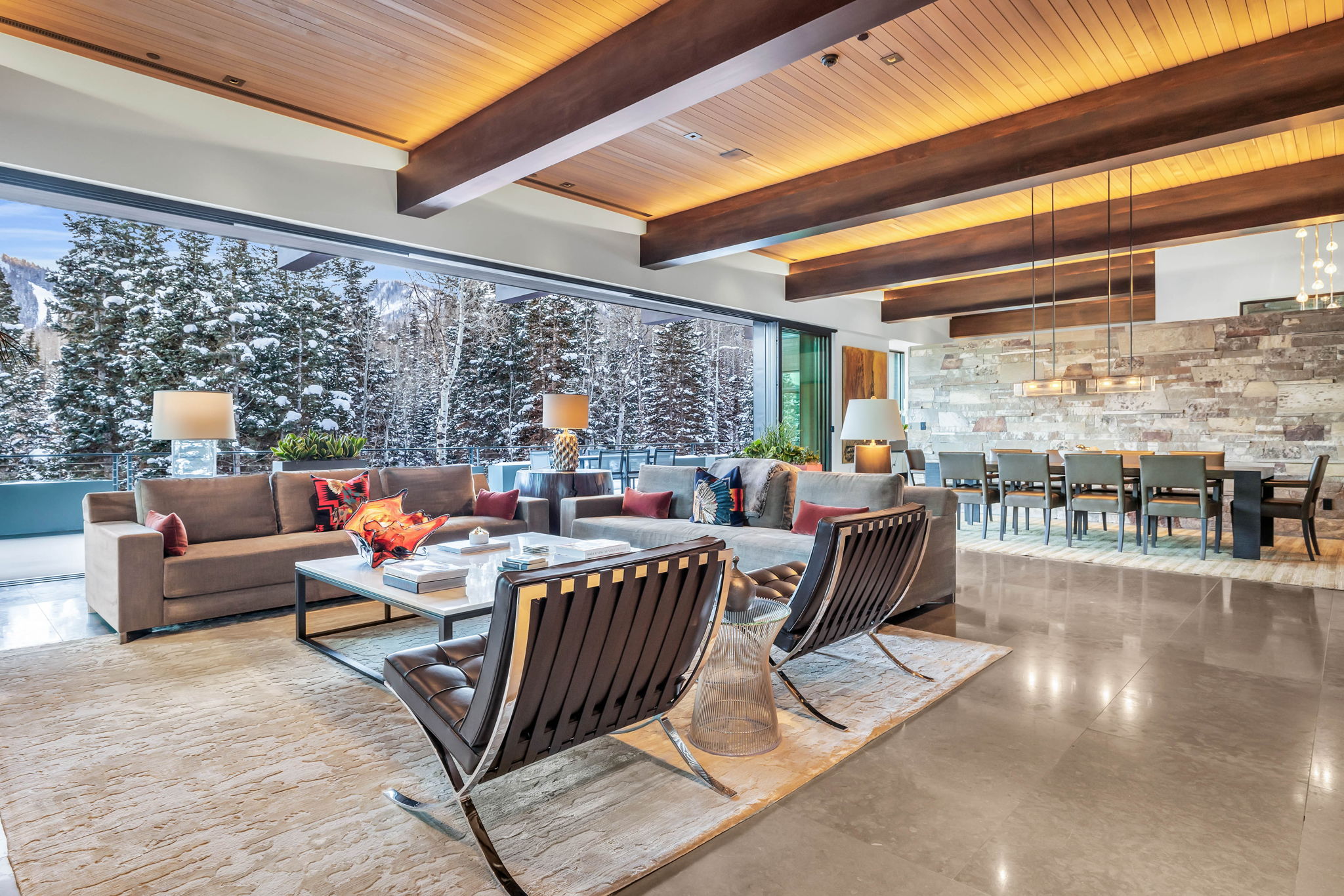 Spacious open-concept living area featuring exposed wood beams, a full-height stone accent wall, and expansive glass doors opening to a snow-covered mountain and forest landscape