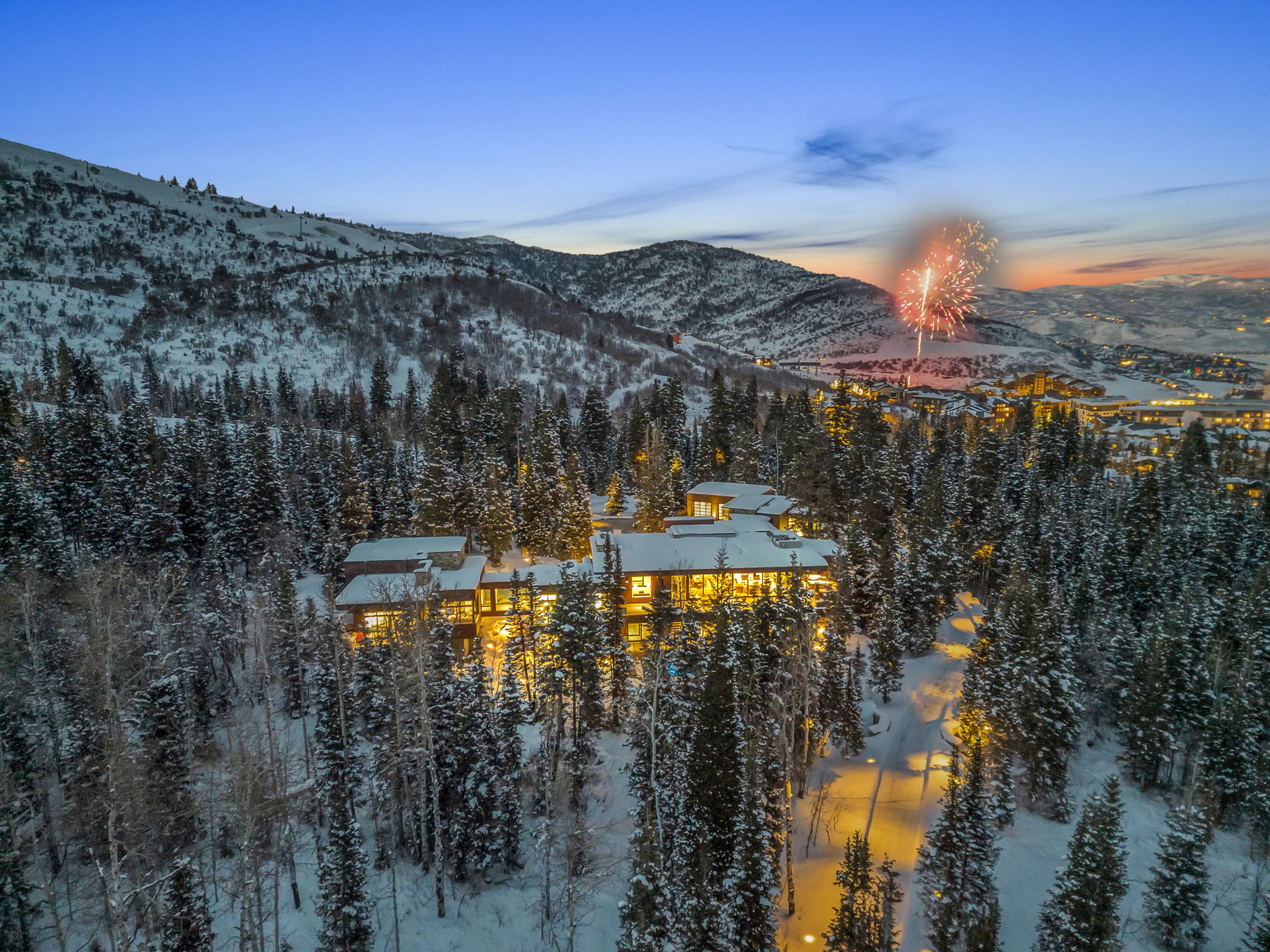 Extensive property nestled among snow-covered evergreen trees, featuring multiple levels, numerous illuminated windows, and a multi-peaked roofline