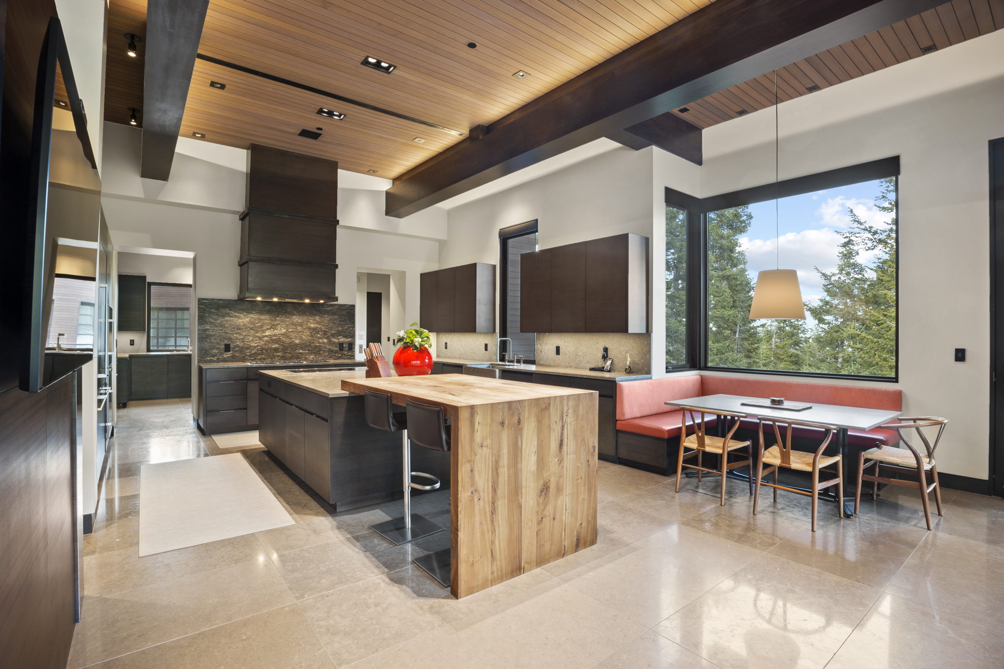 Contemporary kitchen featuring a large central island with a wood-finish breakfast bar, dark cabinetry, a stone-tiled backsplash, and a built-in banquette seating area under expansive corner windows