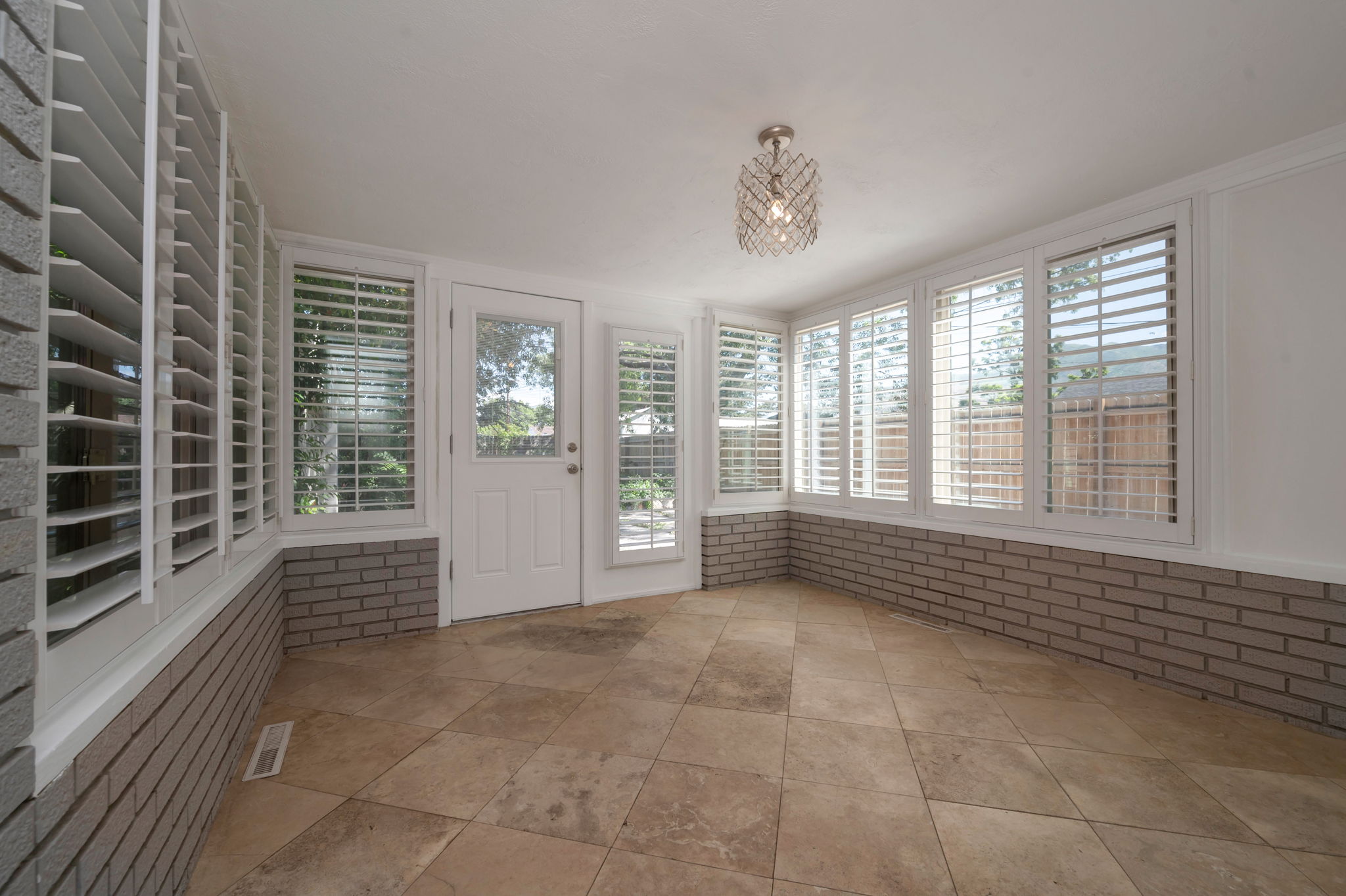 Bedroom 3/Den/Sunroom featuring diagonal tile flooring, a half-wall of brick, multiple windows with plantation shutters, a glass-paned exterior door, and a decorative ceiling light fixture