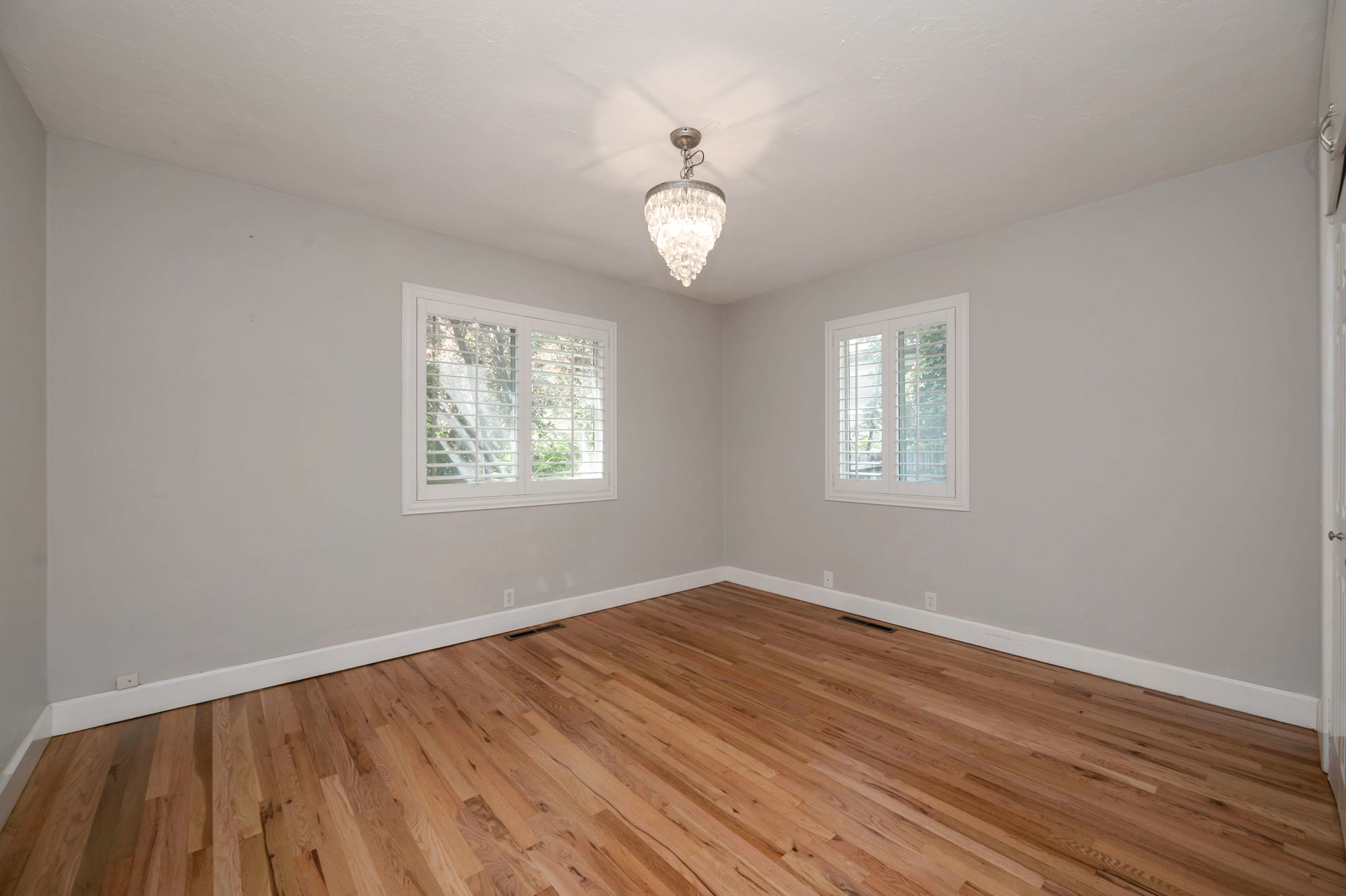 Bedroom 1 featuring wood-finish flooring, two windows with white plantation shutters, white baseboards, and a crystal chandelier