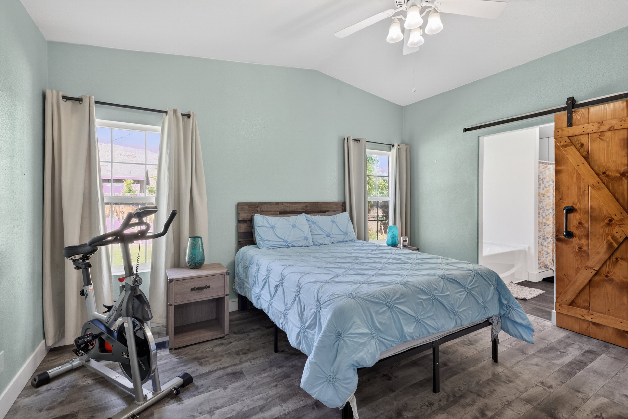 Vaulted ceiling with a ceiling fan light fixture, dual windows with curtain rods, wood-finish flooring, and a rustic barn door with black hardware