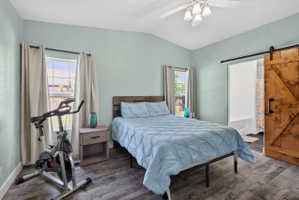 Vaulted ceiling with a ceiling fan light fixture, dual windows with curtain rods, wood-finish flooring, and a rustic barn door with black hardware