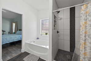 Bathroom featuring a corner soaking tub with a glass block window, dark wood-finish flooring, a shower with white subway tile, a dark hexagon tile accent strip, and a dark hexagon tile shower floor