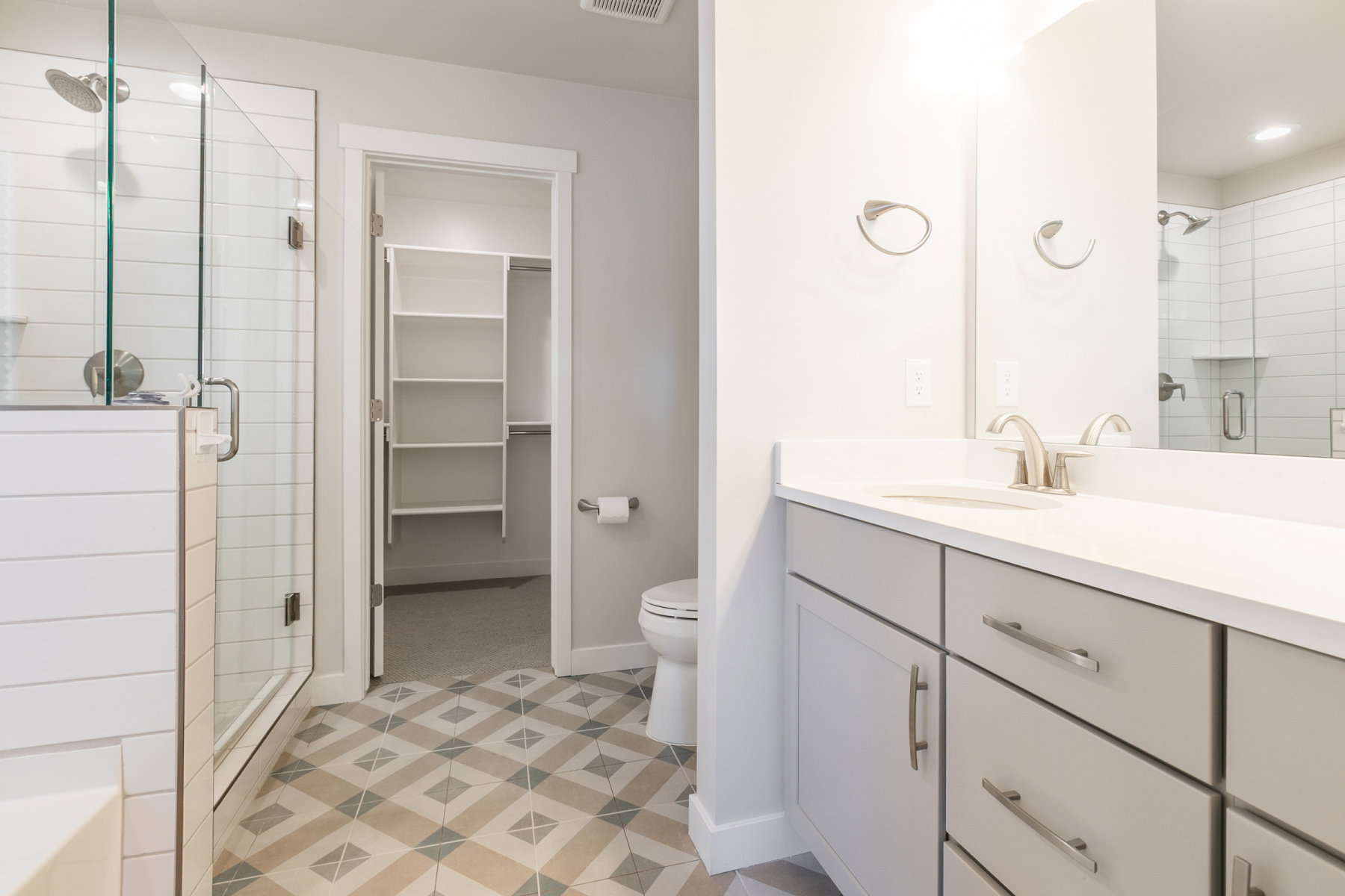 Bathroom featuring patterned floor tiling, a glass-enclosed shower with white subway tile, a light gray vanity with a white countertop and brushed nickel hardware, and an adjacent walk-in closet with built-in shelving