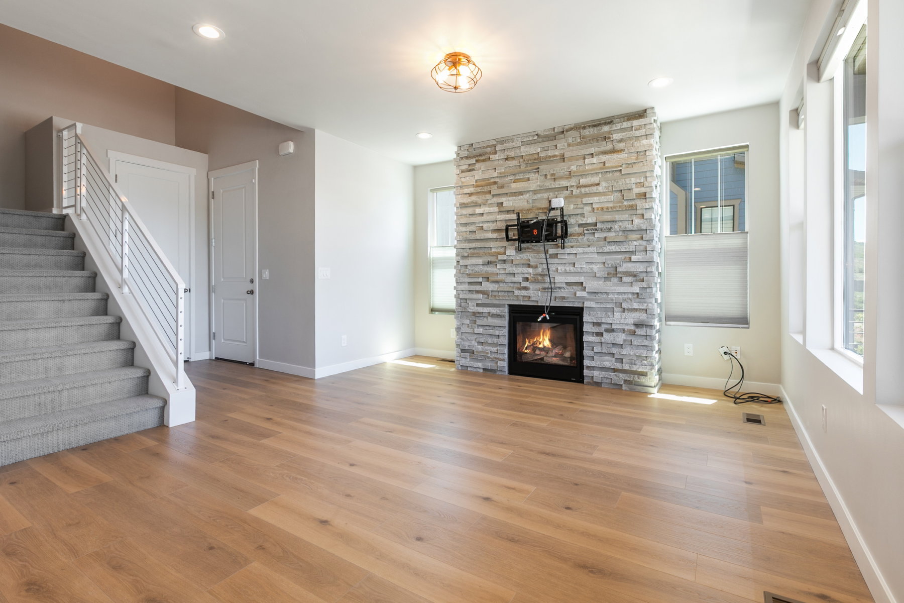 Spacious living area featuring a stacked stone fireplace with a black insert, wood-finish flooring, multiple windows, and a carpeted staircase with a modern metal railing