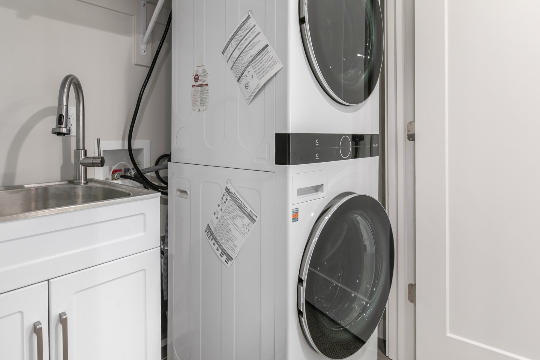 Utility area featuring a stainless steel sink with a high-arc faucet, white base cabinetry with brushed nickel hardware, and a stacked front-load washer and dryer