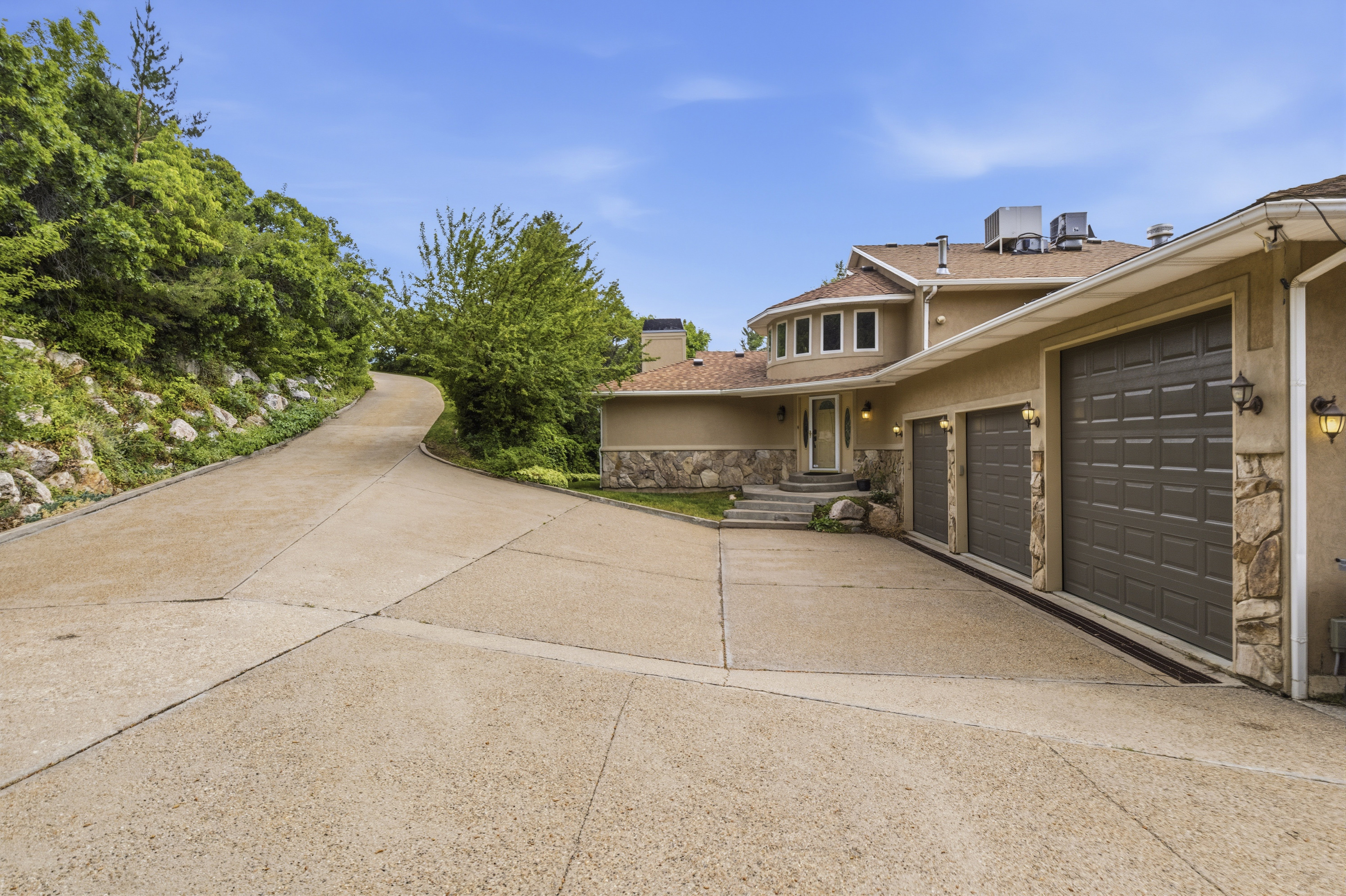 Expansive concrete driveway featuring a three-car garage with individual doors, a stucco exterior with stone accents, and a winding approach surrounded by mature greenery