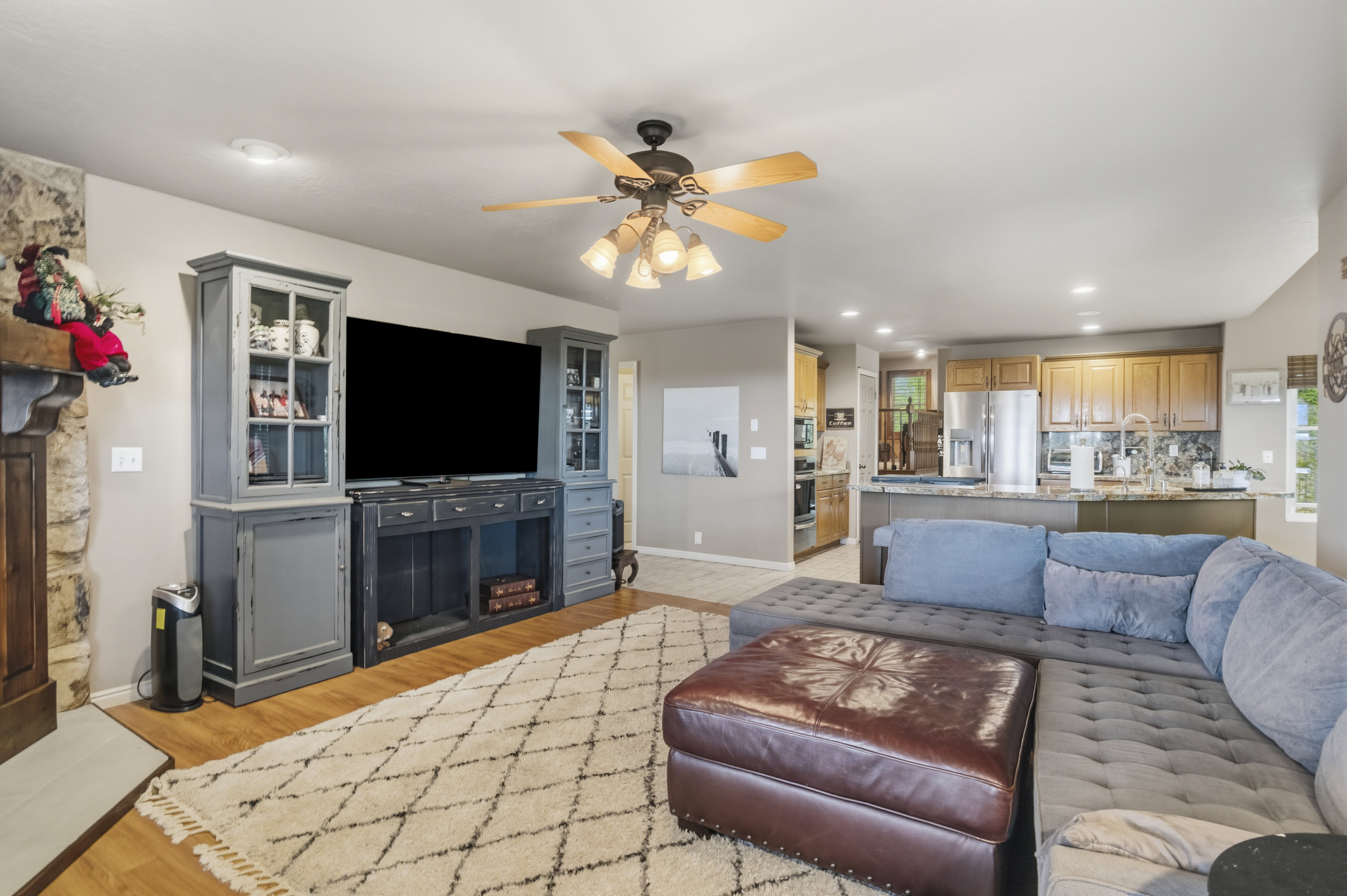Open-concept living space featuring wood-finish flooring, a stone fireplace, and a ceiling fan with integrated lighting