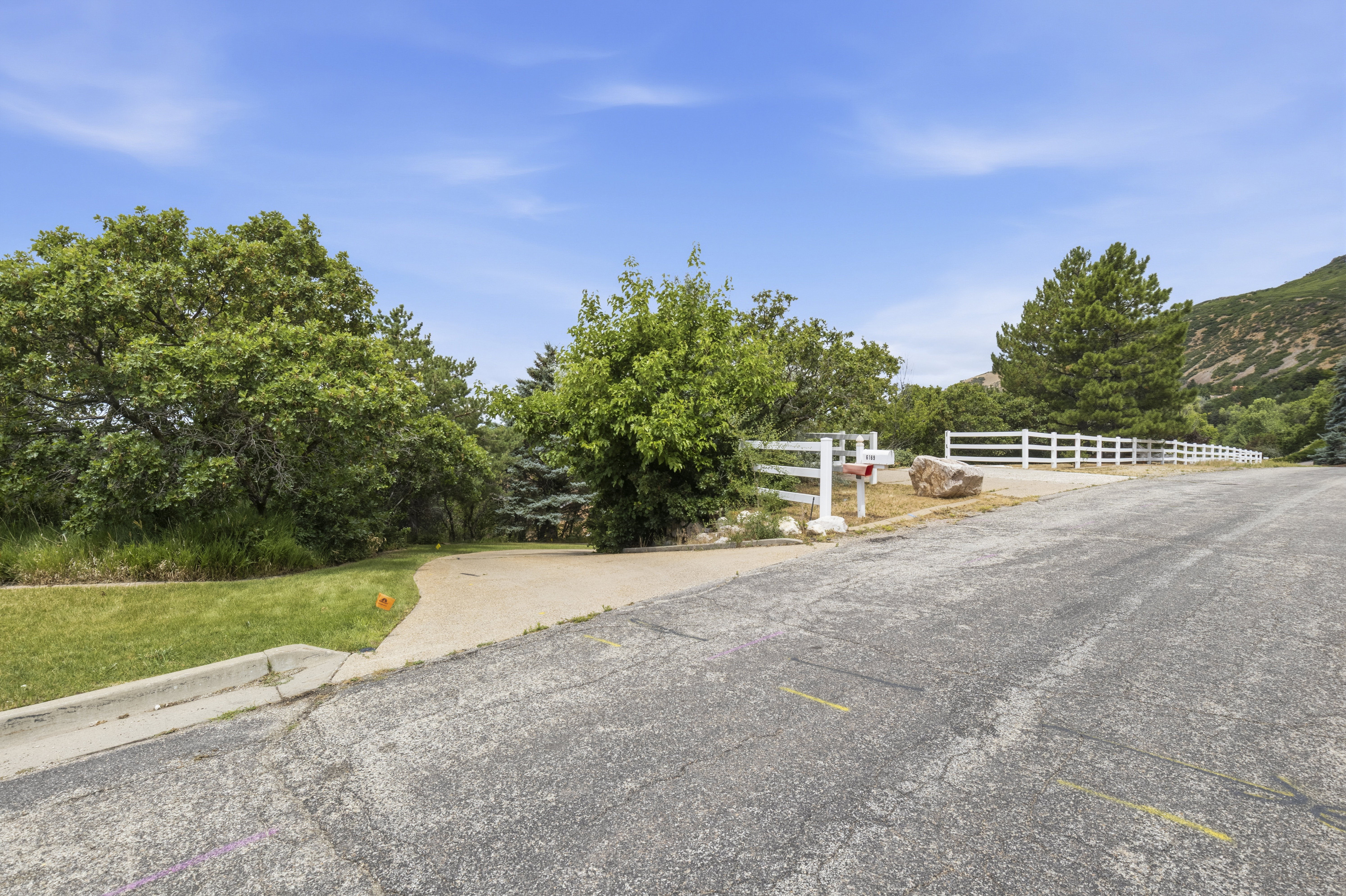 Paved driveway entrance with a white picket fence, mature trees, and a mountain backdrop