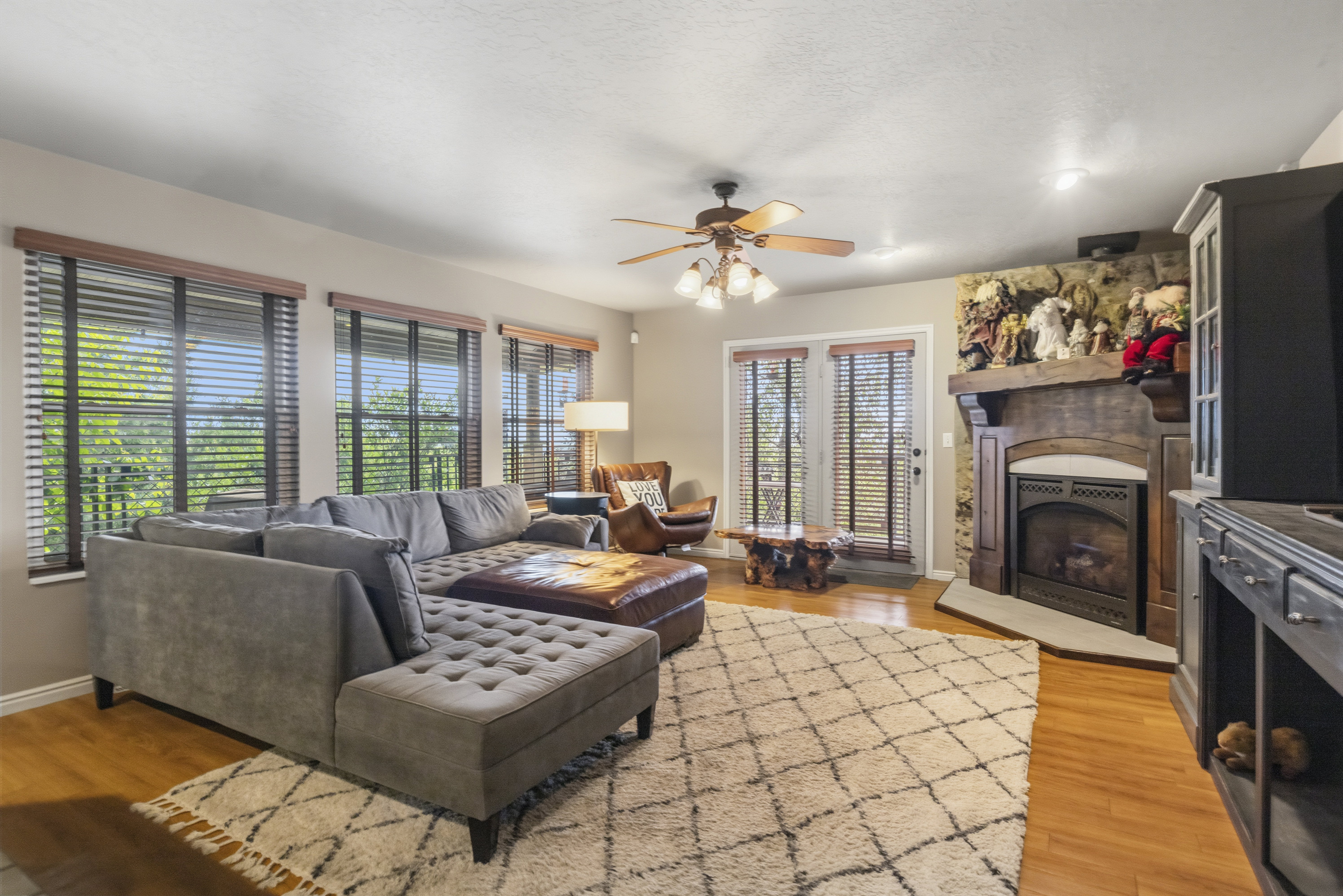 Living area featuring wood-finish flooring, a stone-accented fireplace with a wood mantle, a ceiling fan with integrated lighting, and multiple windows with horizontal blinds