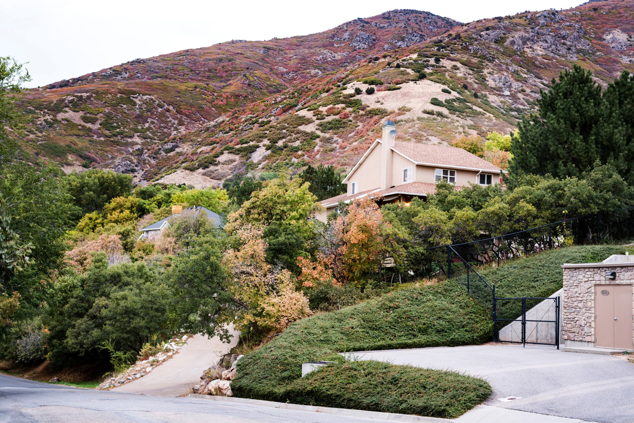 Sloped property featuring a stucco-sided residence, a stone-clad utility structure, a paved driveway, and mature trees against a mountainous backdrop