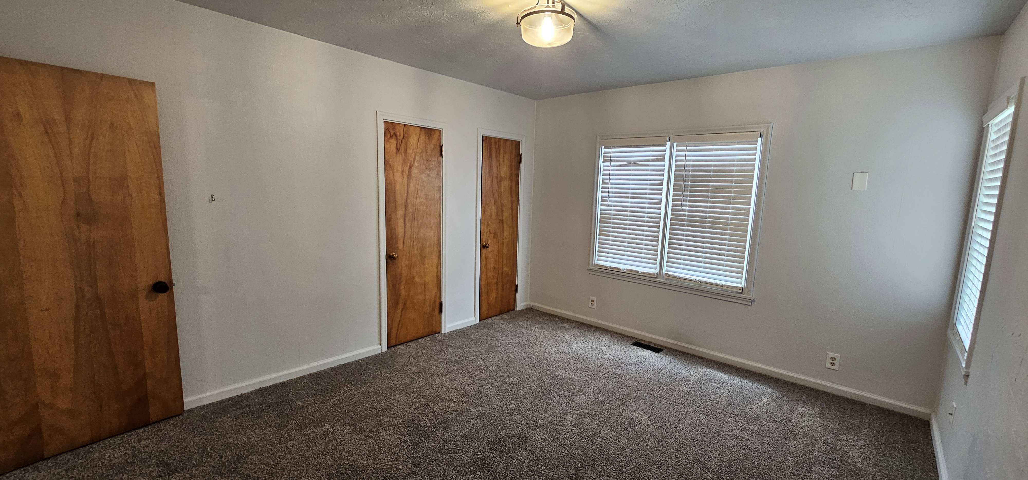 Carpeted interior space featuring white walls, two wood-finish doors, and two windows with blinds