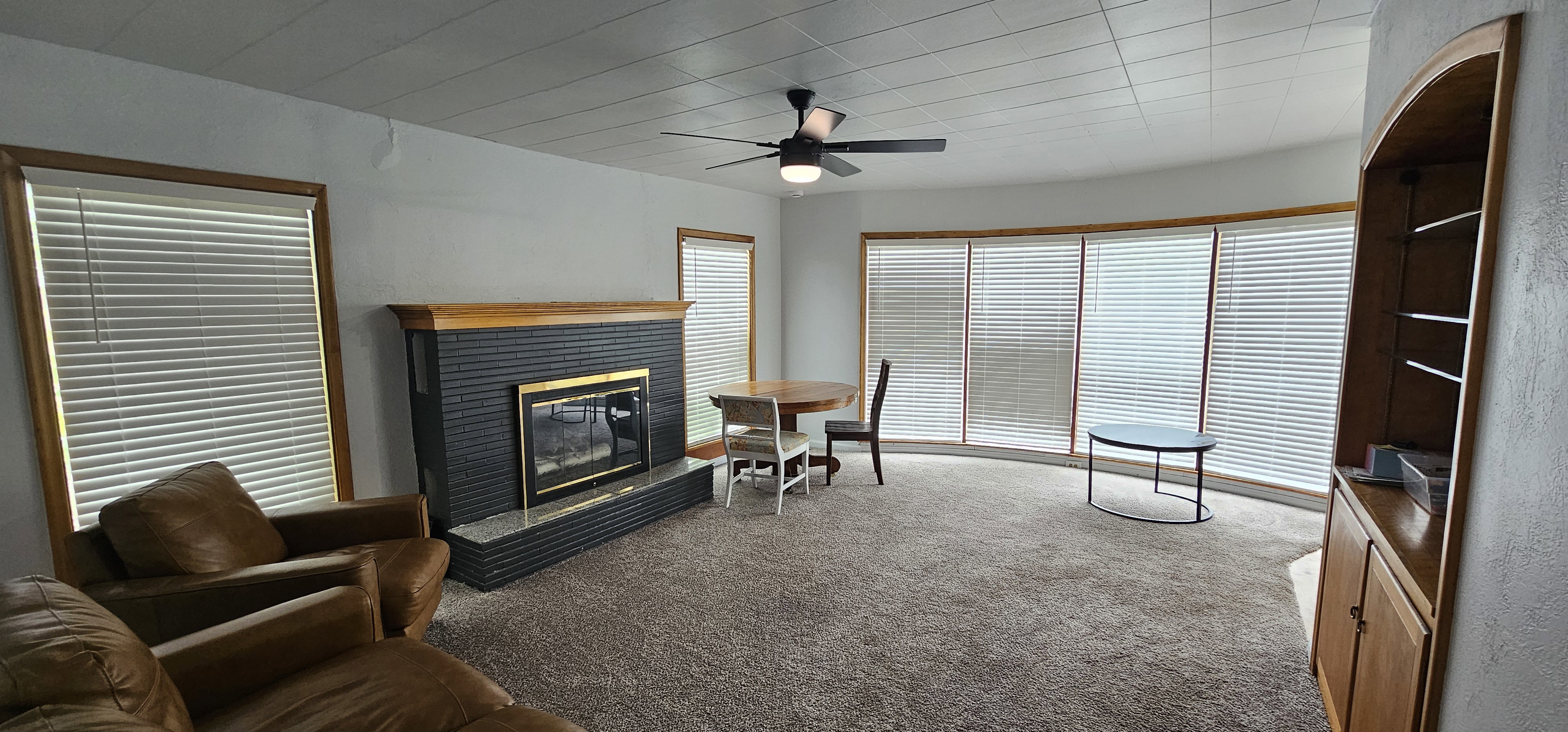 Living area featuring a textured fireplace with a wood mantle, a ceiling fan, and multiple windows with blinds