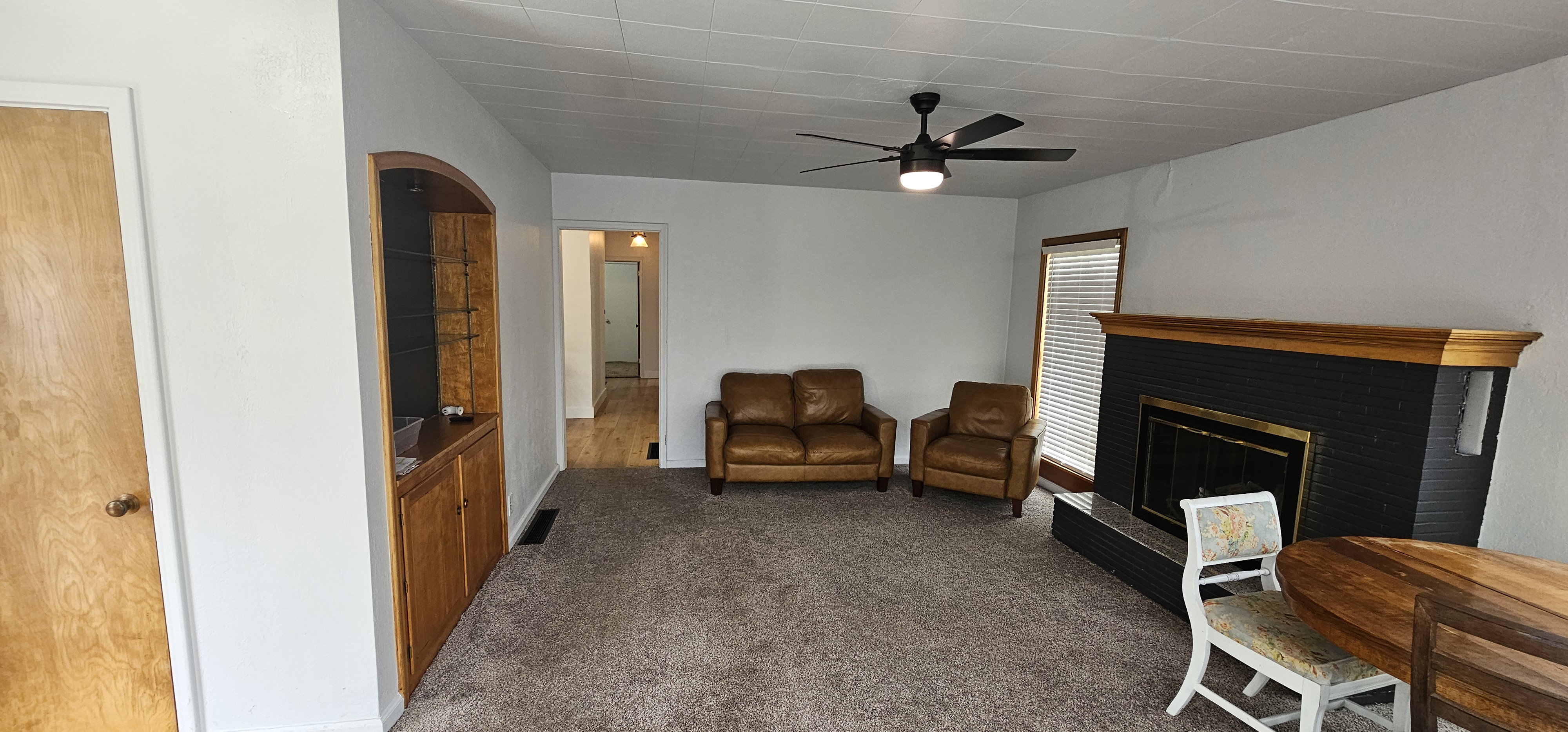 Spacious living area featuring a fireplace with a black surround and wood mantel, neutral carpeting, and a ceiling fan with integrated lighting