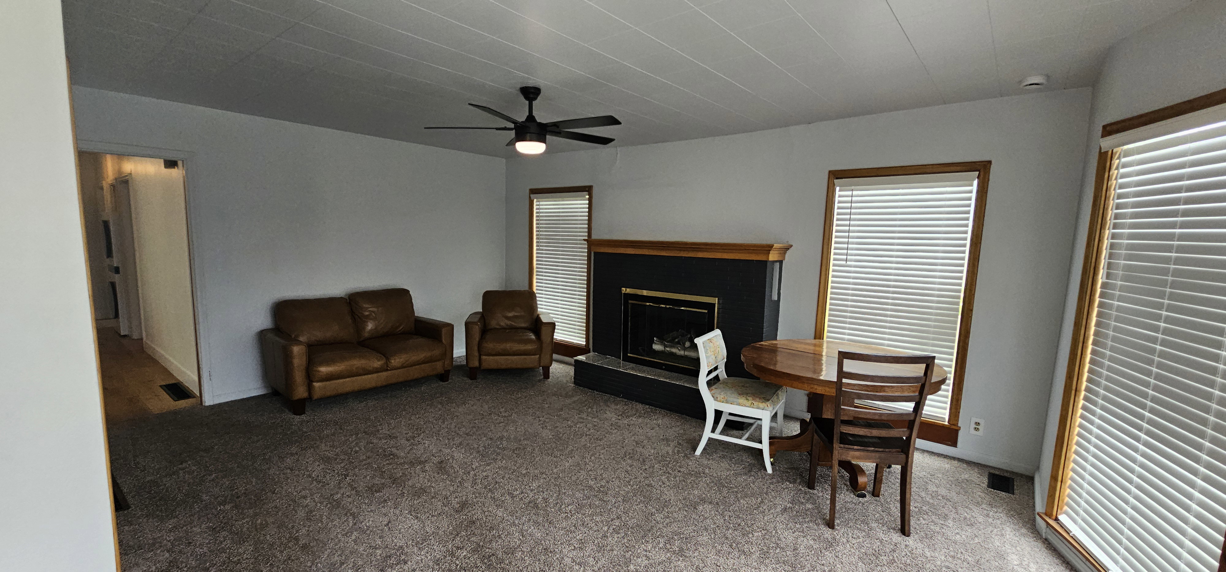Living area featuring a central fireplace with a dark surround and wood mantel, neutral-toned carpeting, and a ceiling fan with integrated lighting