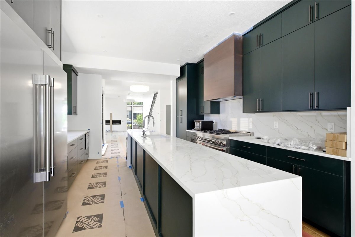 Modern kitchen featuring a waterfall island with a stainless steel sink, dark green cabinetry, white stone countertops, and a full-height stone backsplash