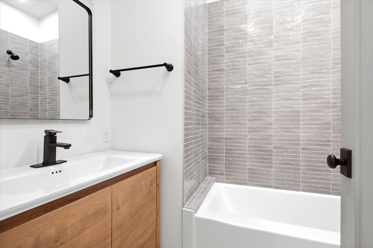 Bathroom featuring a white bathtub with light gray horizontal tile surround, a wood-finish vanity with an integrated white sink, and matte black fixtures