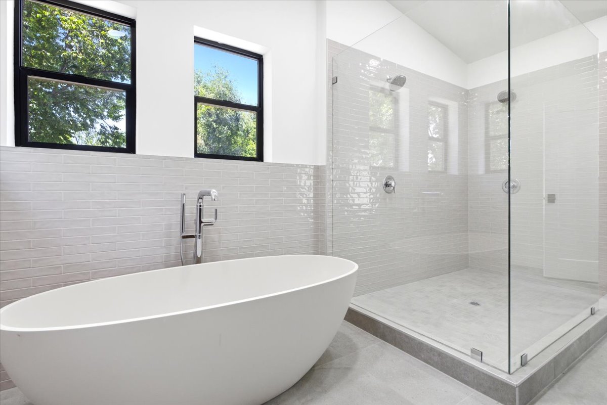 Bathroom featuring a freestanding oval soaking tub with floor-mounted faucet, a spacious glass-enclosed shower with dual showerheads, light-toned horizontal tile work, and two rectangular windows