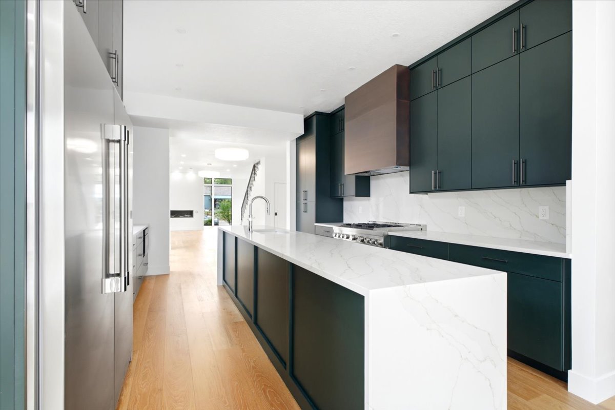 Contemporary kitchen featuring a large waterfall island with a sink, dark green cabinetry, stainless steel appliances, a copper-tone range hood, and light wood-finish flooring