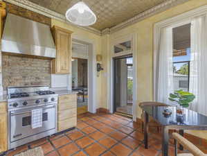 Kitchen featuring terra cotta flooring, stainless steel range with matching hood, brick-look backsplash, light wood cabinetry, and a pressed tin ceiling