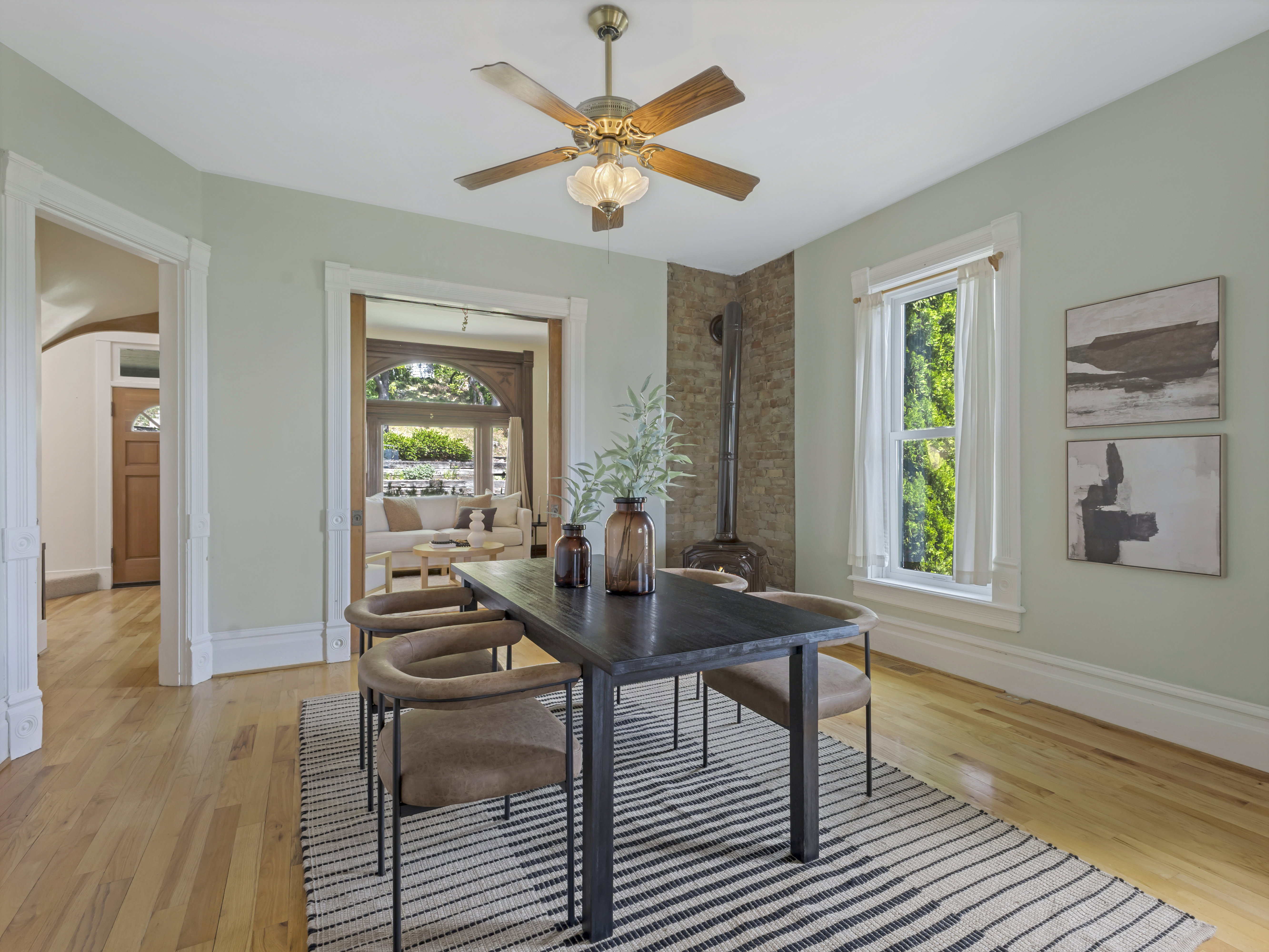 Spacious room featuring wood-finish flooring, a ceiling fan with integrated lighting, and a brick-accented wall with a wood-burning stove