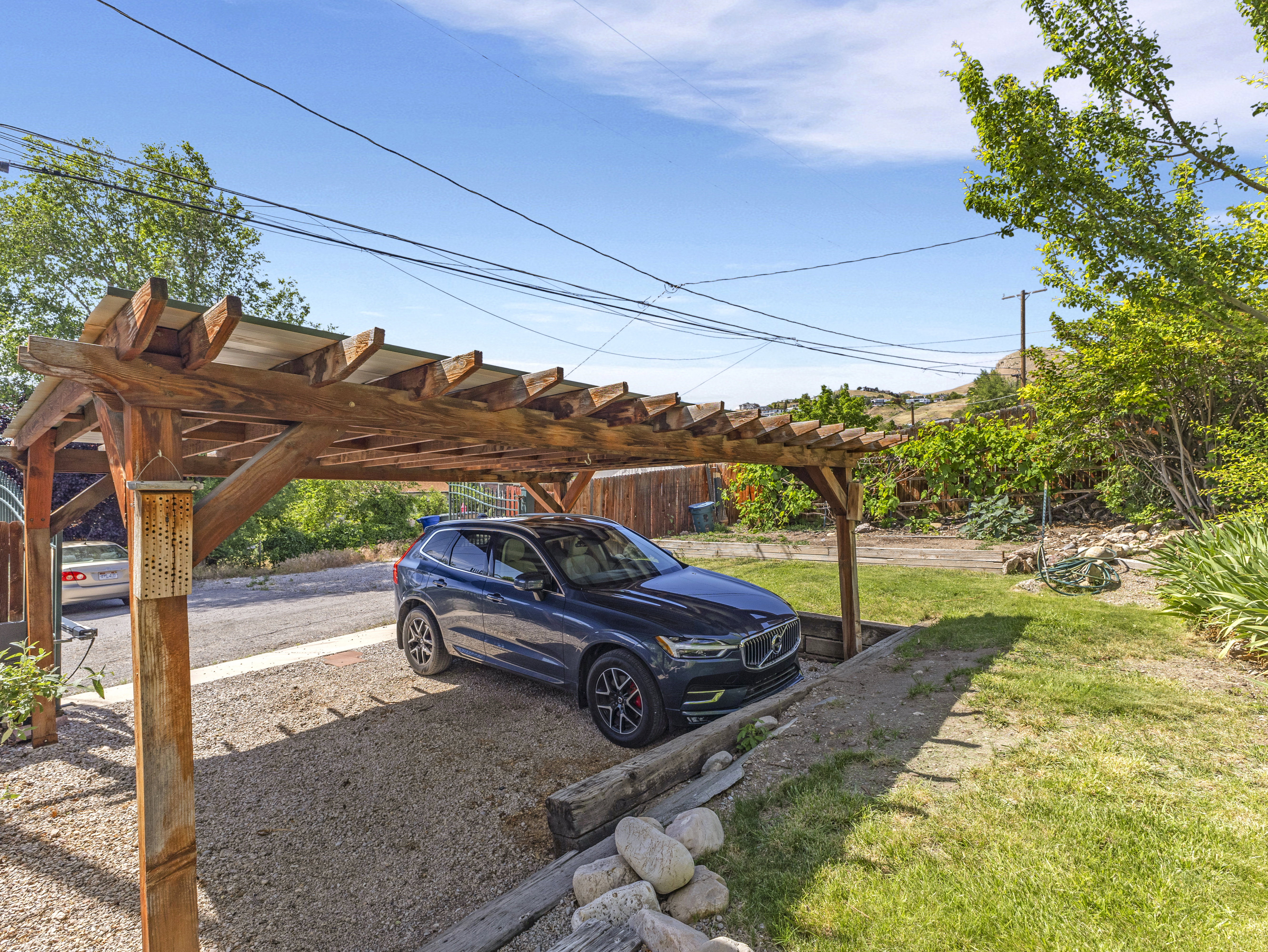 Wood pergola with a translucent roof over a gravel parking area, bordered by a retaining wall and green lawn