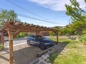 Wood pergola with a translucent roof over a gravel parking area, bordered by a retaining wall and green lawn