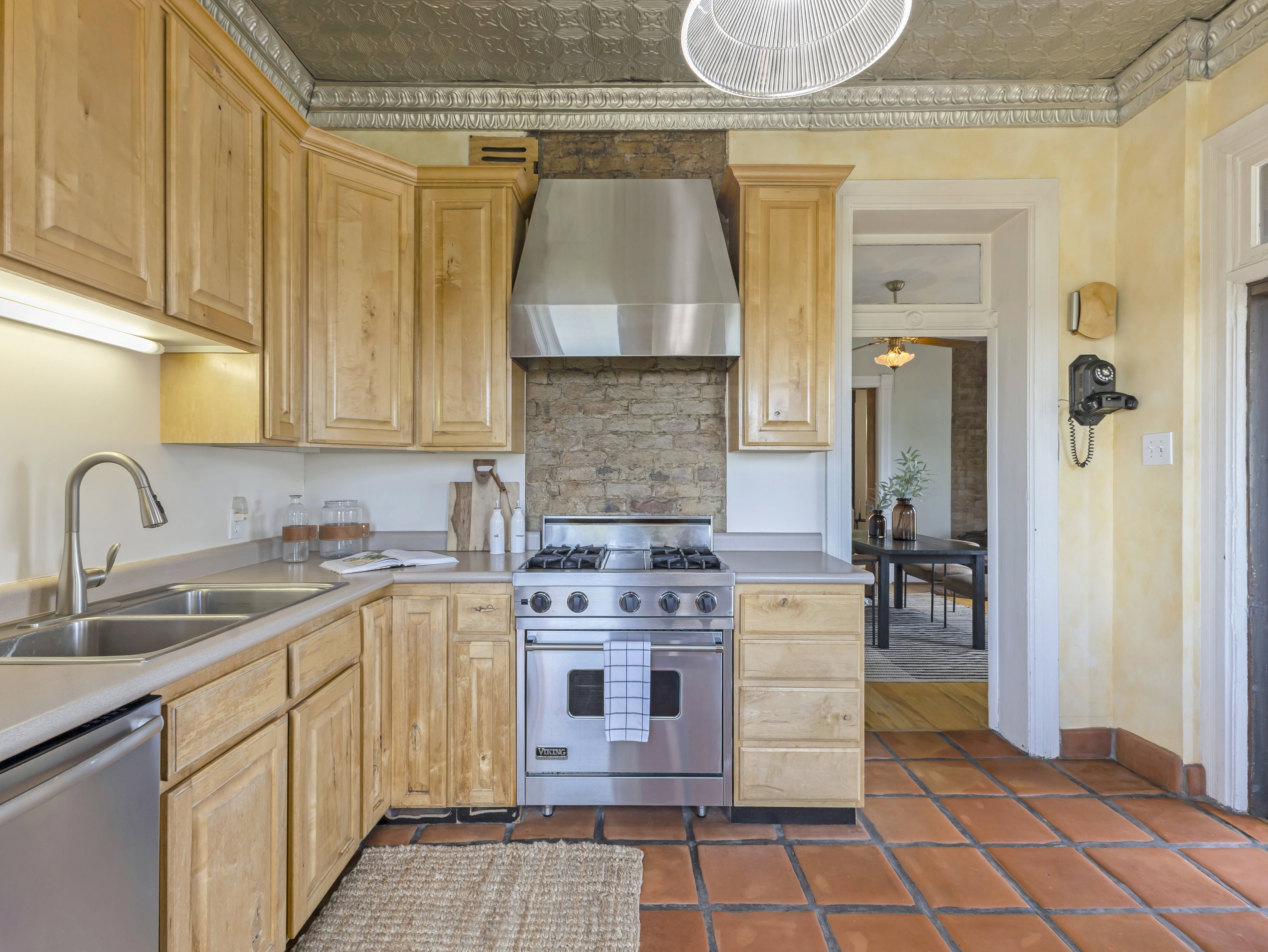 Kitchen featuring terra cotta tile flooring, light wood cabinetry, stainless steel range with hood, double basin sink, and a tin ceiling with decorative trim