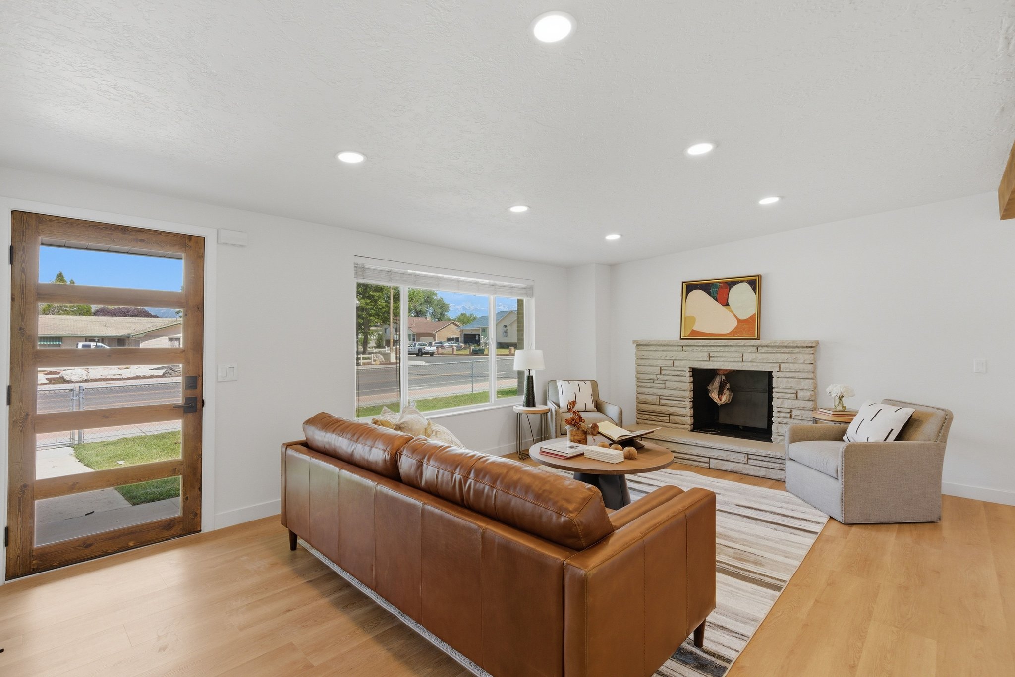 Bright living area featuring wood-finish flooring, a contemporary wood-paneled door with horizontal glass inserts, recessed lighting, a stone-faced fireplace, and large windows