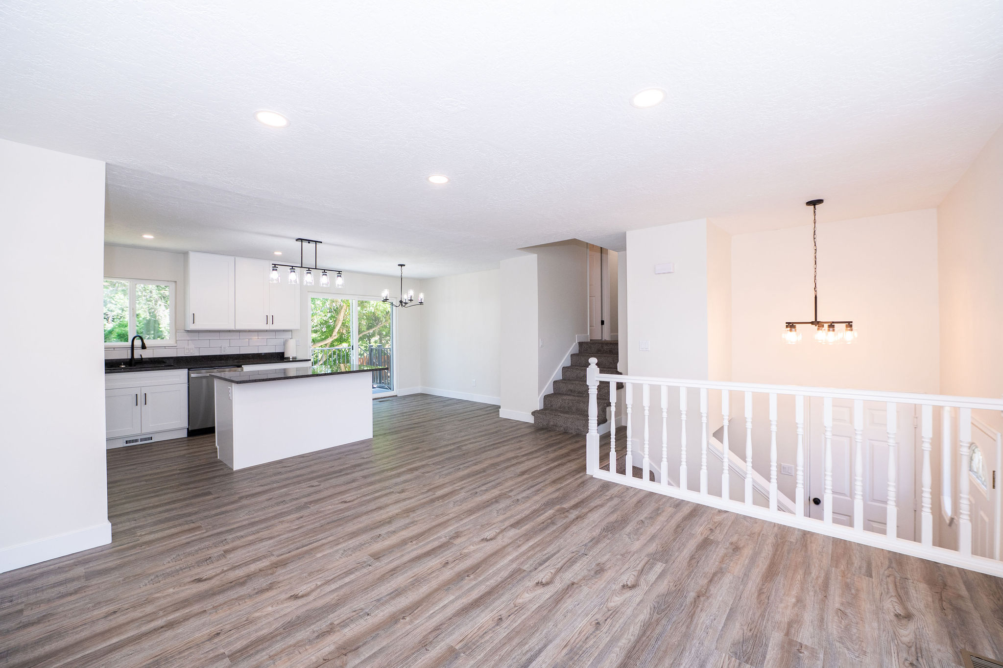 Open-concept living area featuring wood-finish flooring, a contemporary kitchen with white cabinetry and dark countertops, a kitchen island, and a white baluster railing